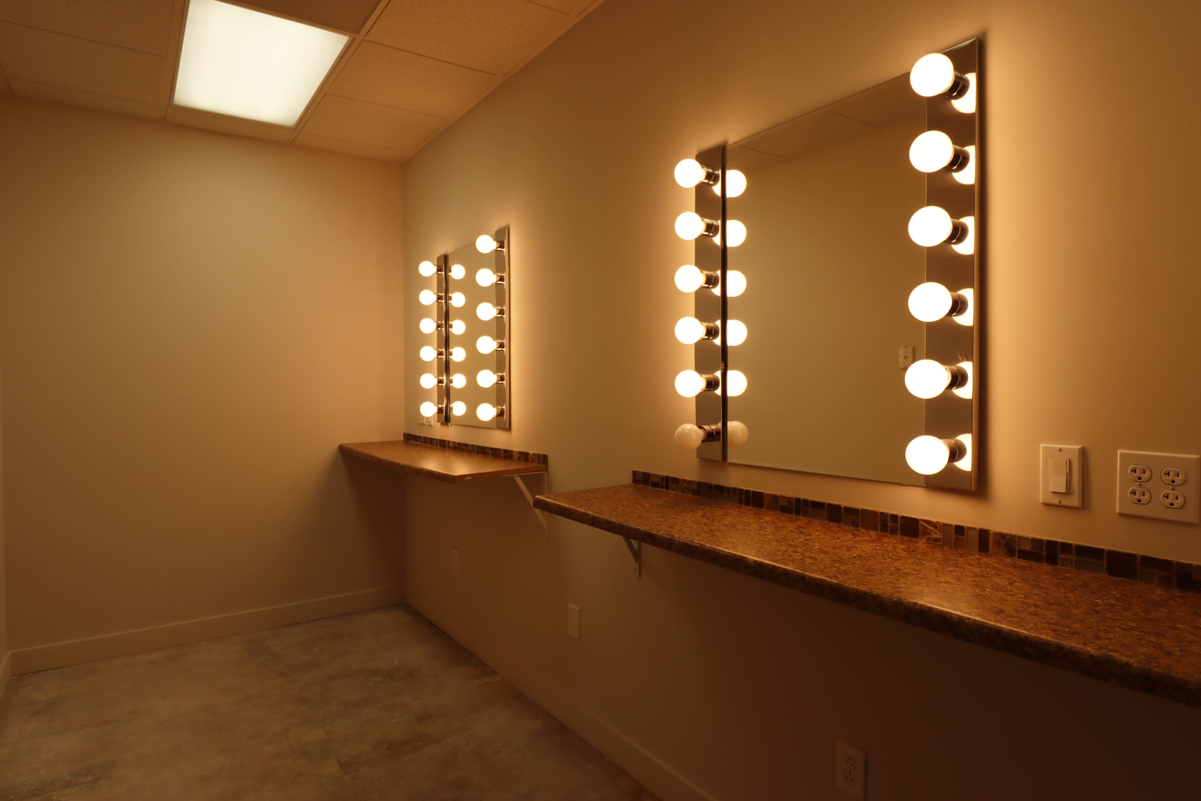 The image shows a well-lit vanity area with two mirrors framed by round bulbs and matching countertop surfaces.