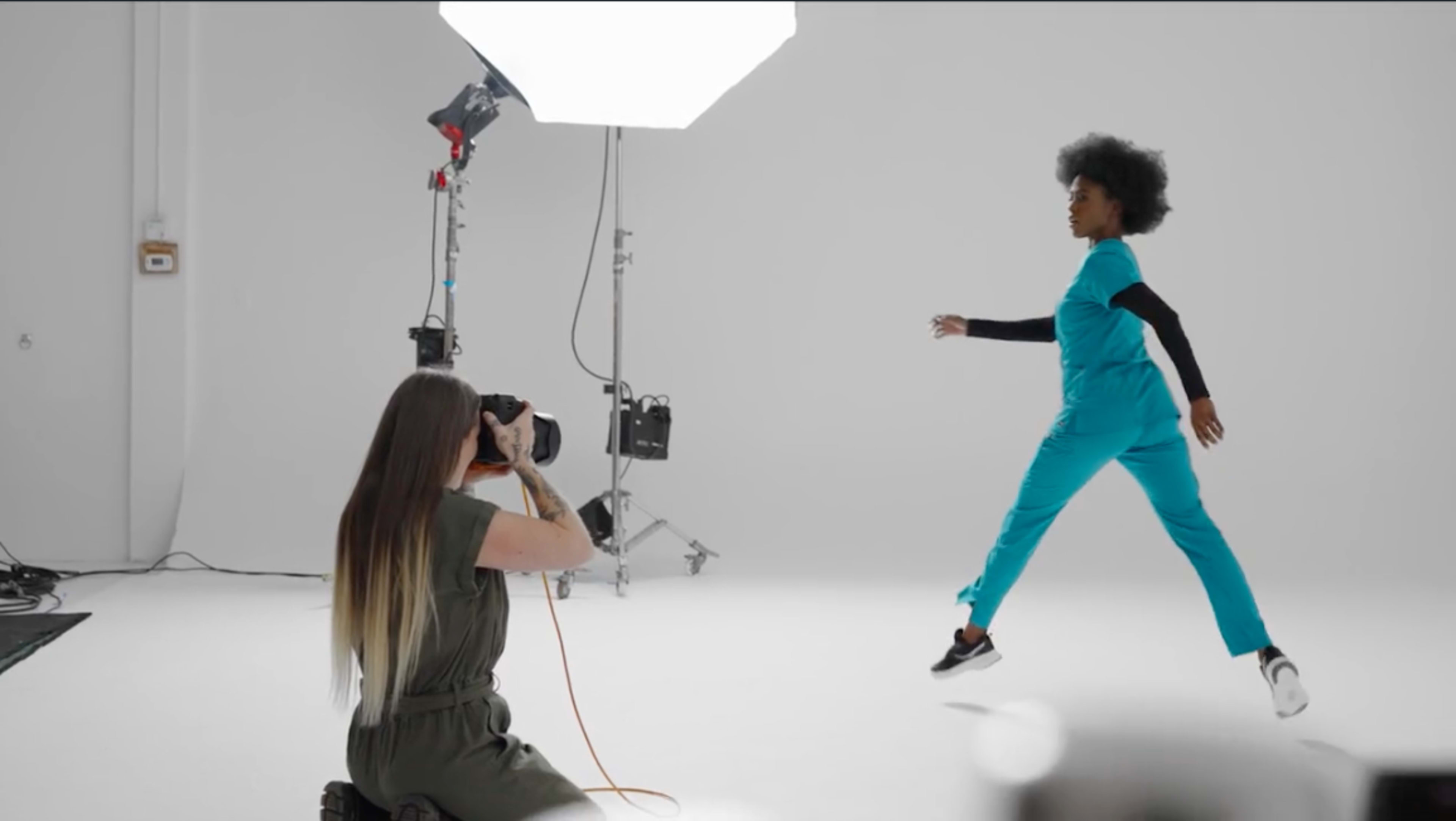 A photographer captures a model in mid-air while she poses in a studio with a white background, wearing turquoise scrubs.