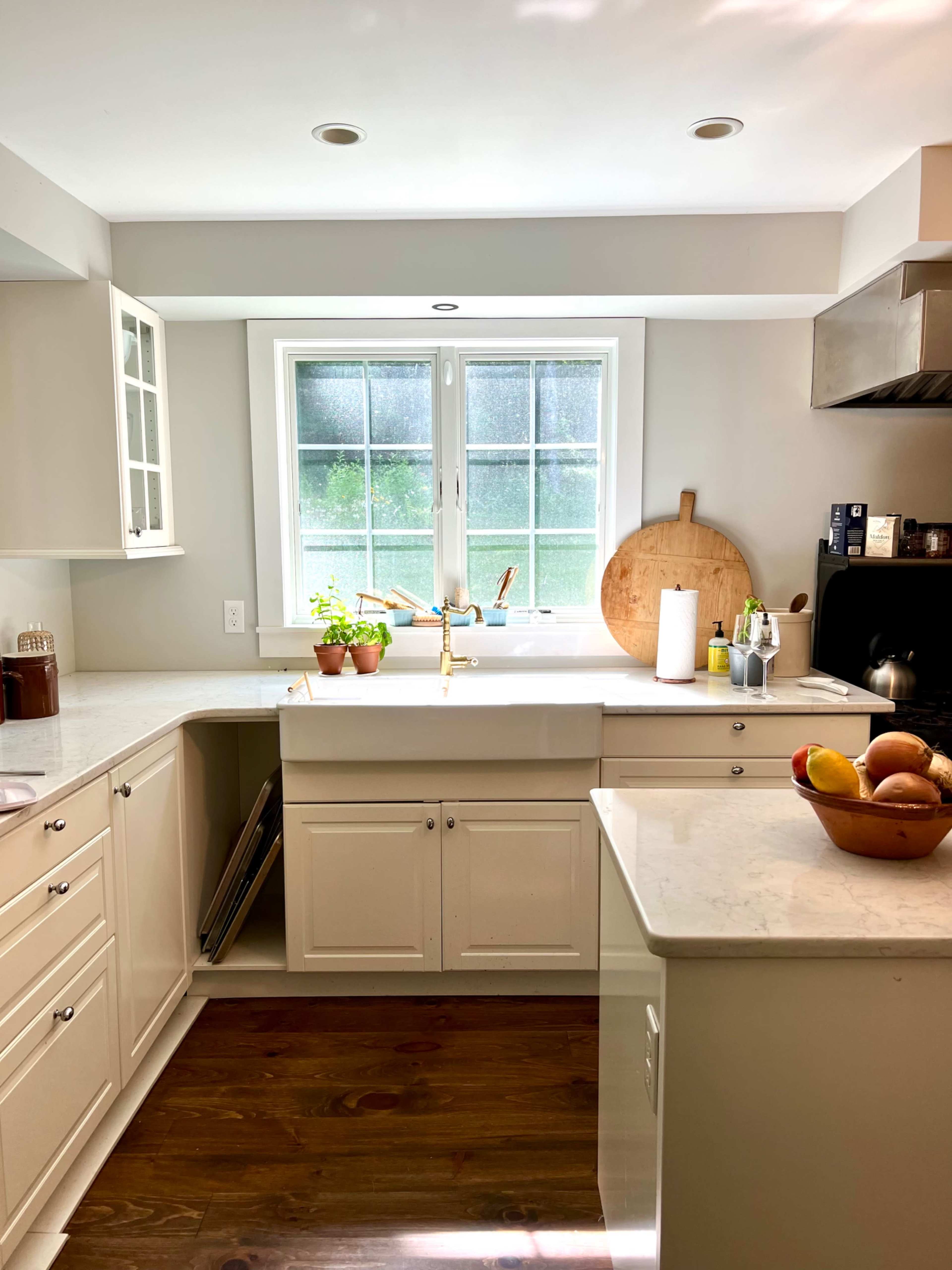 A bright kitchen features white cabinetry, a large window above the sink, and potted plants on the countertop.