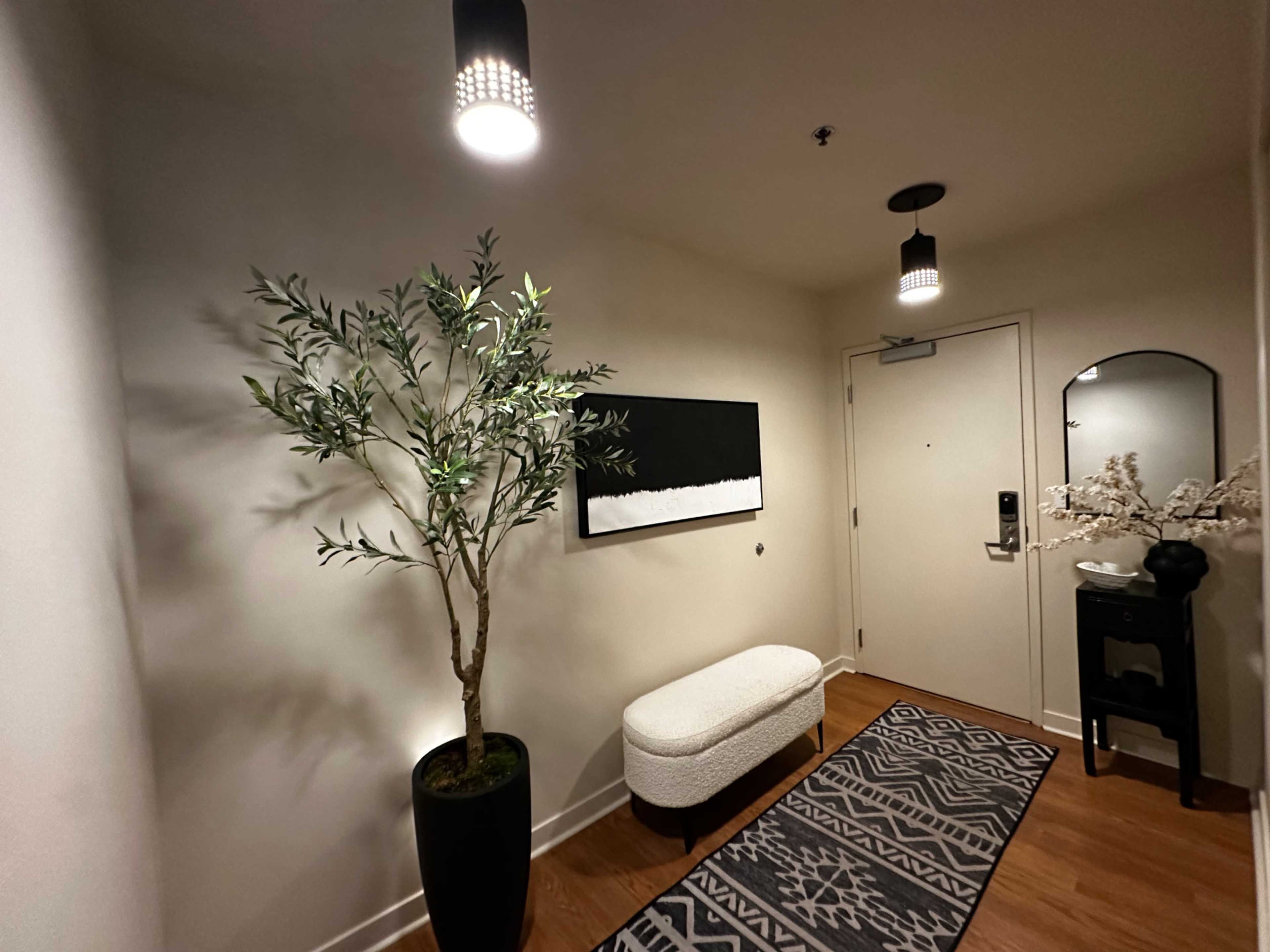 The image shows a narrow hallway featuring a potted plant, a bench, a piece of wall art, and a door with a lock.
