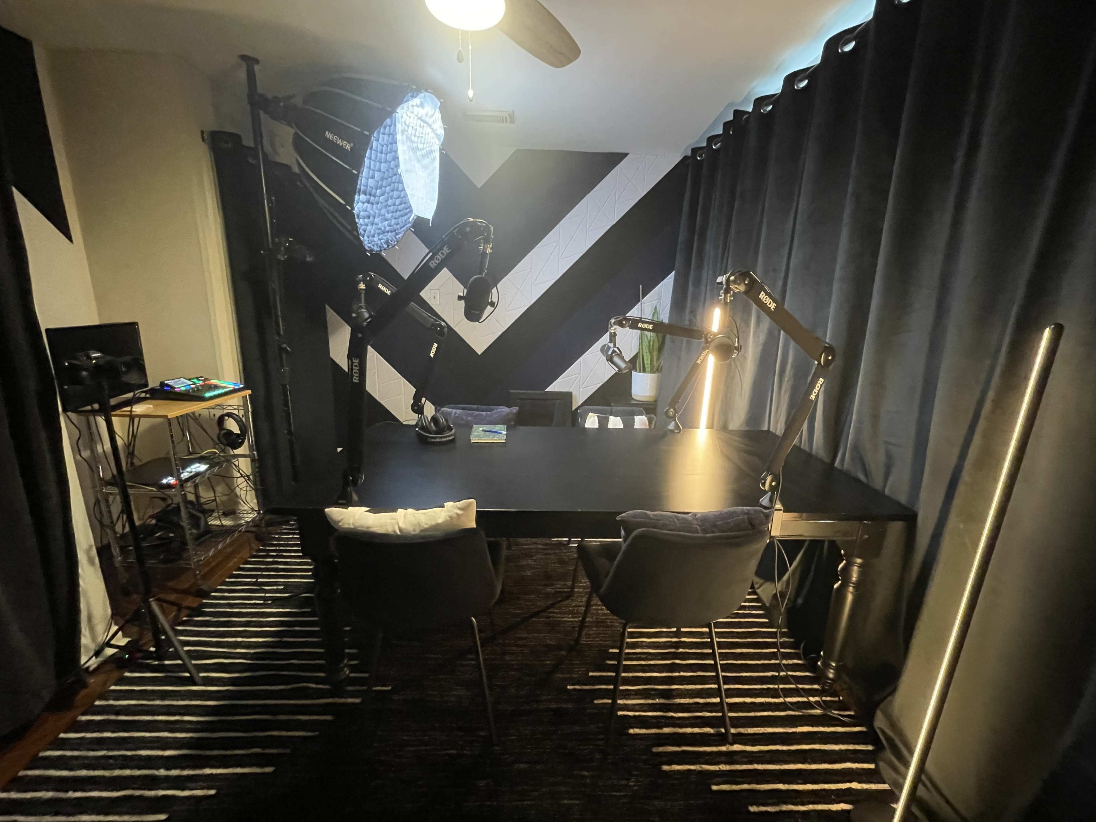 The image shows a room with a dark striped wall, a large black table equipped with adjustable microphone stands, and two chairs, illuminated by soft lighting and a ceiling fan.