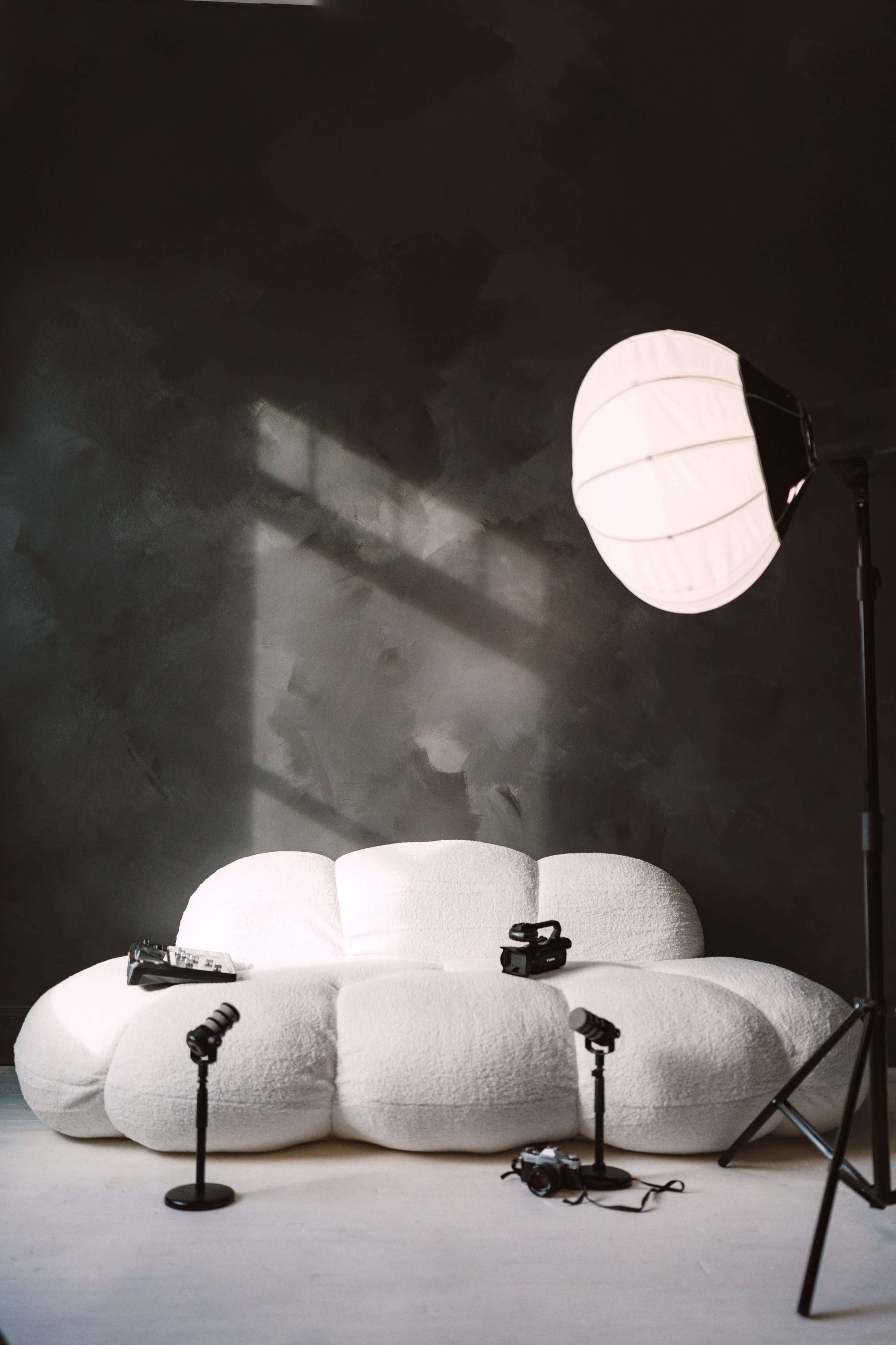 A white, plush couch is positioned against a dark wall, with studio lighting equipment set up nearby.