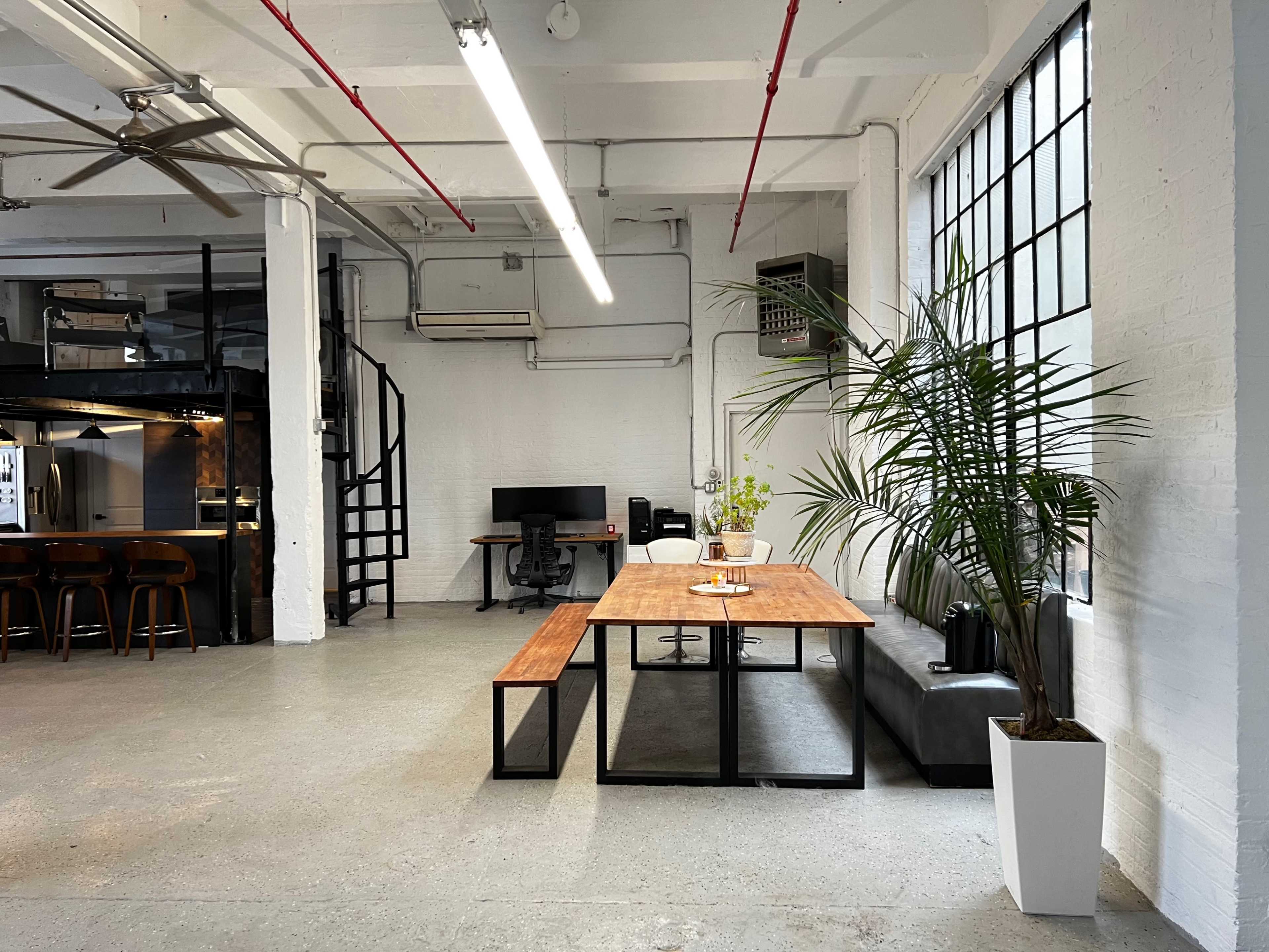 A modern office space featuring a wooden table with benches, a black leather sofa, a computer workstation, and large windows allowing natural light.
