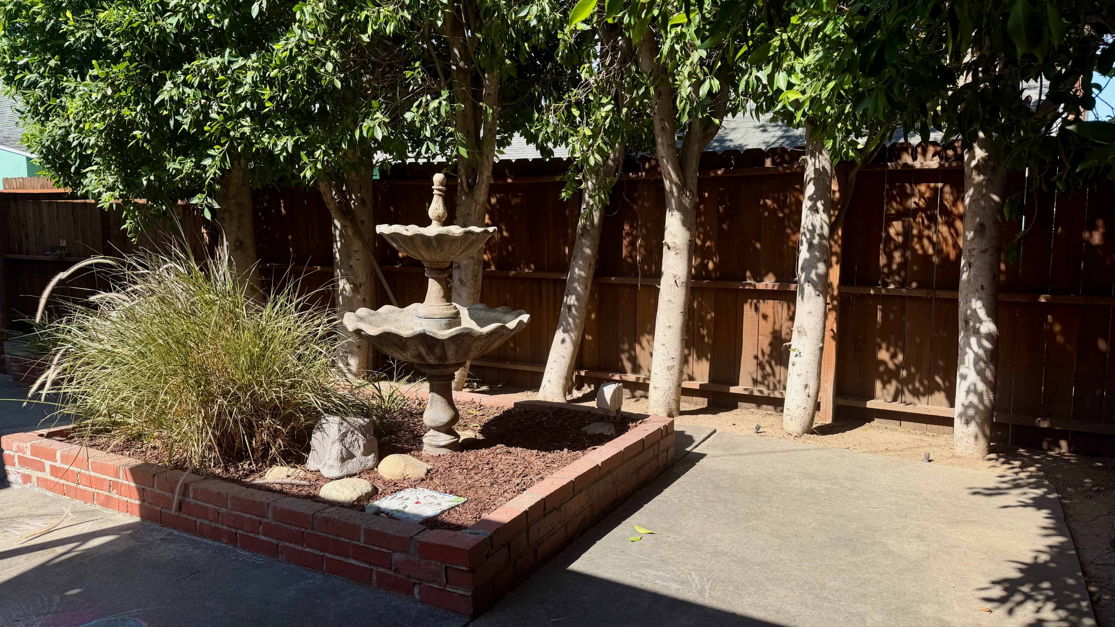 The image shows a backyard area featuring a stone fountain surrounded by landscaping, including plants and a brick border.