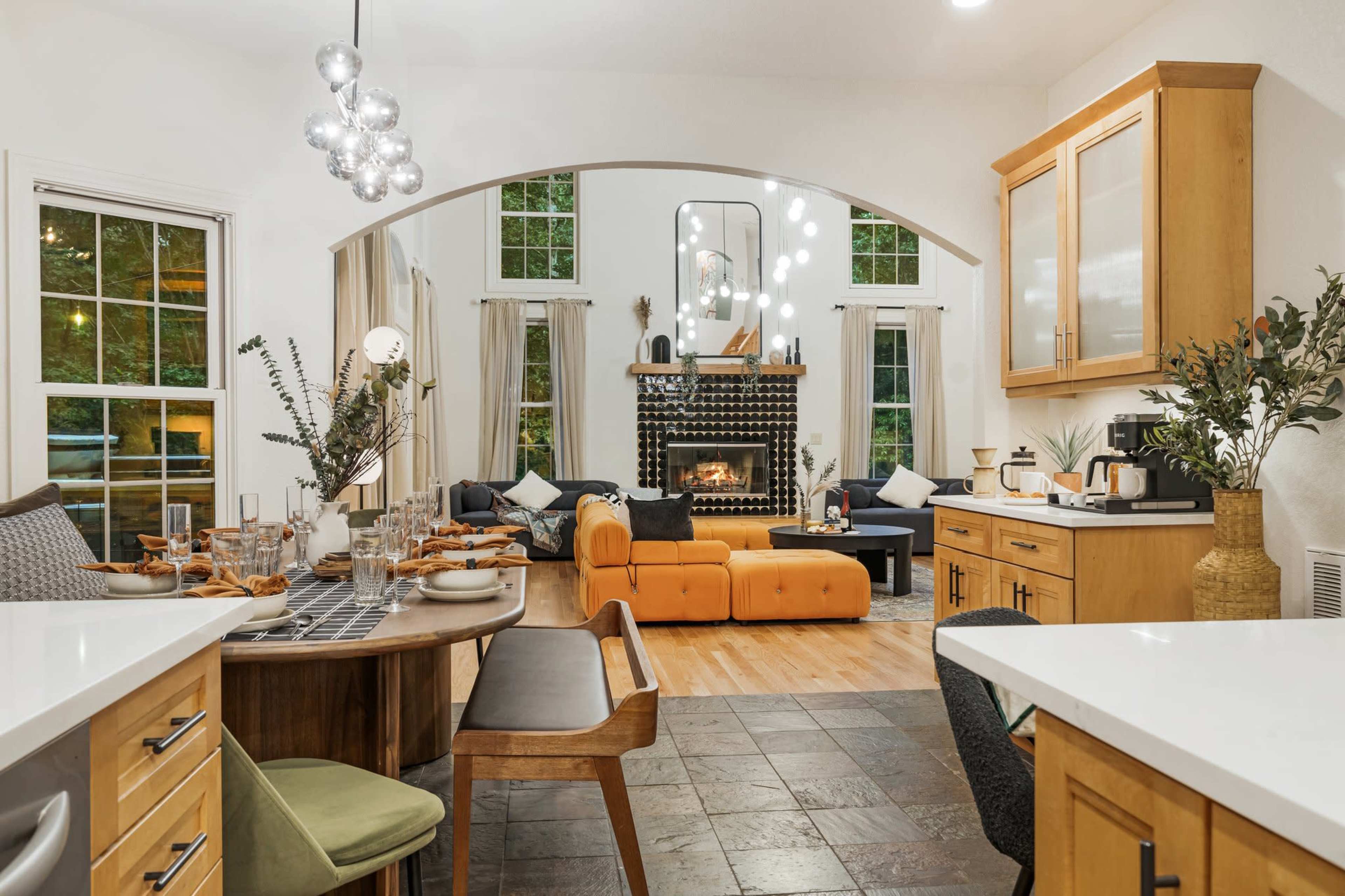 A modern kitchen opens up to a cozy living area featuring a fireplace, large windows, and contemporary furniture.
