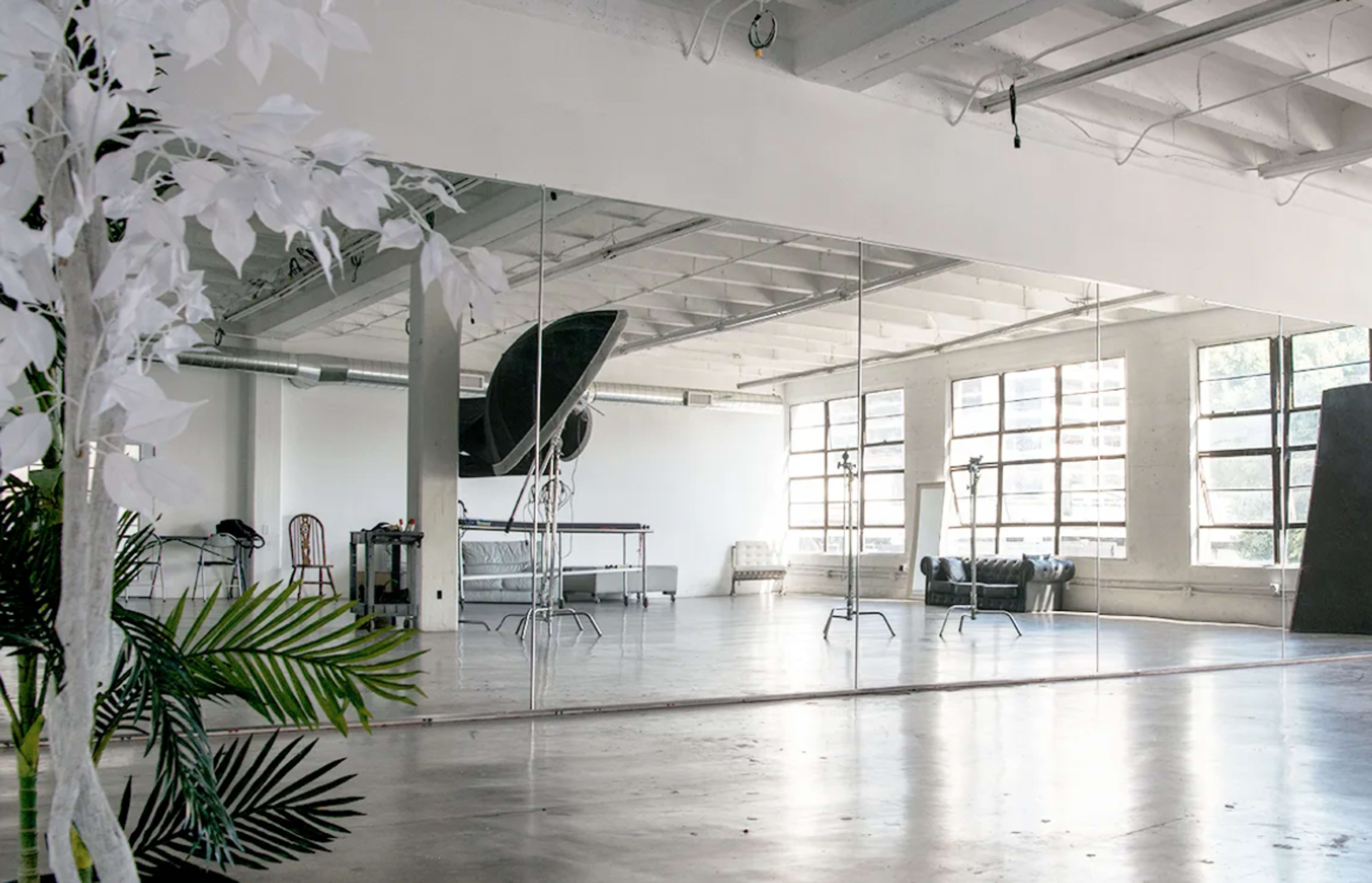 A spacious studio with large windows, a mirrored wall, and minimalist furnishings, including a couch and a chair.