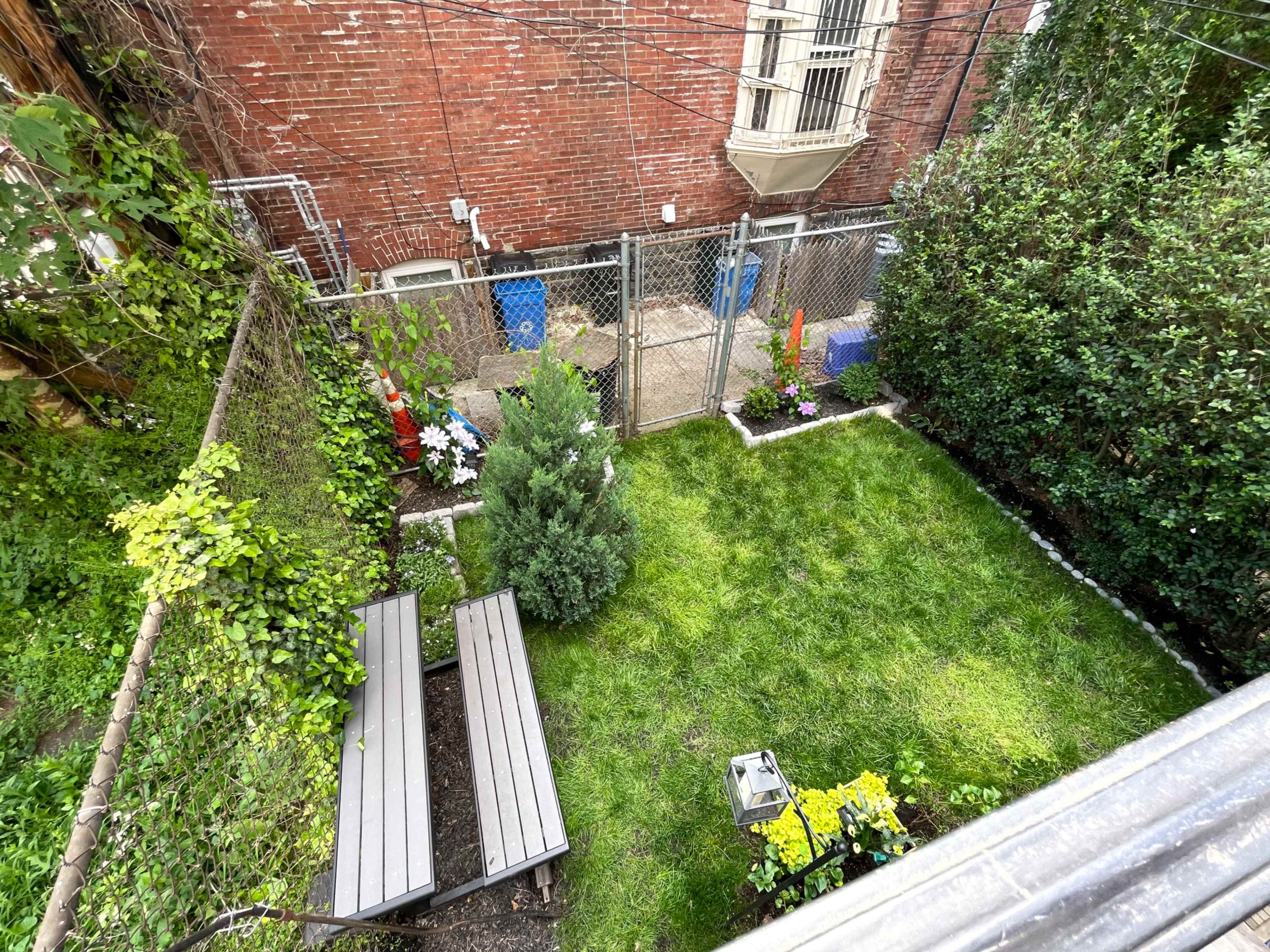 A fenced backyard with a manicured lawn, contains two wooden benches, and is bordered by shrubs and flowers.