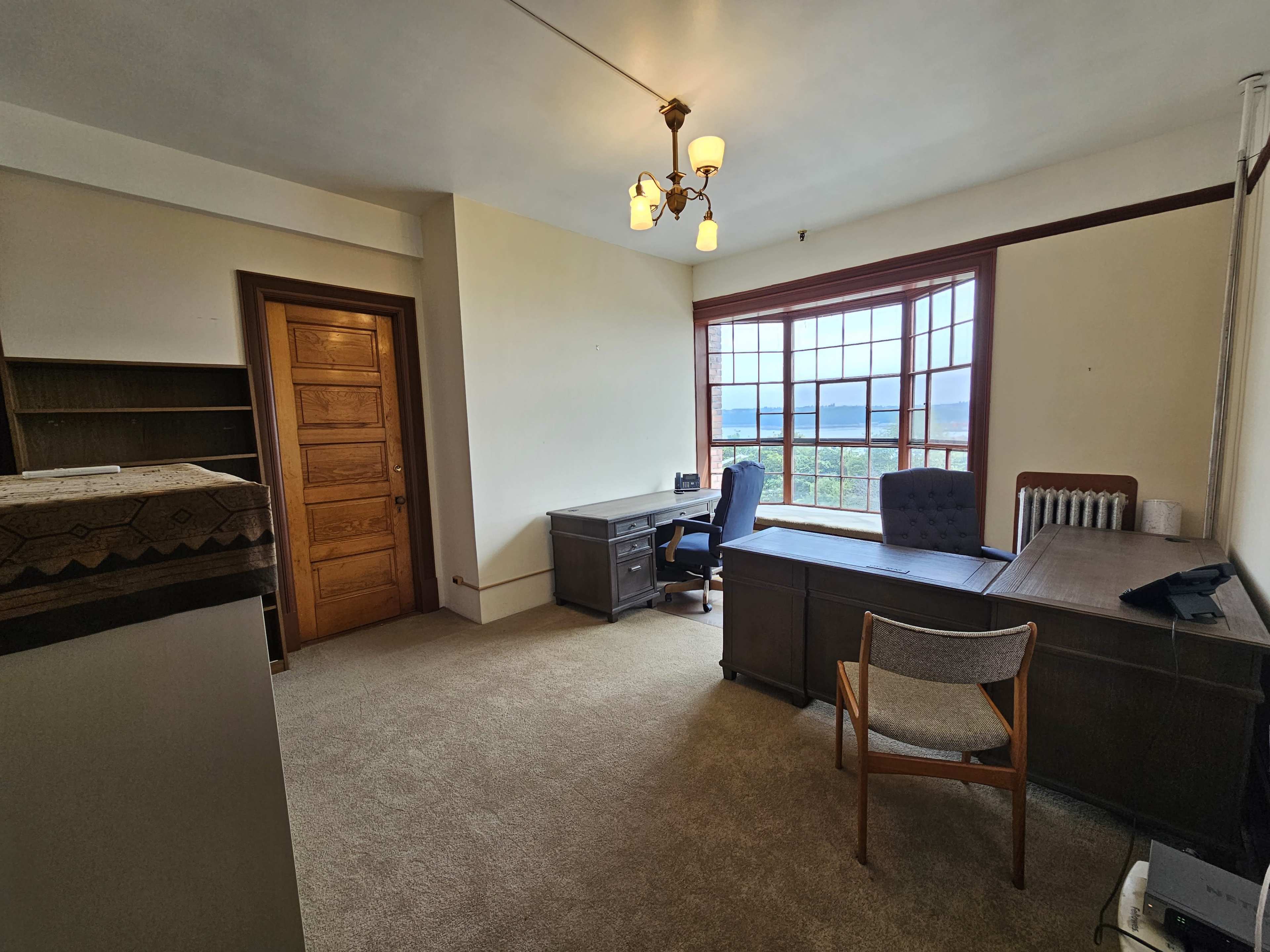 A simple office space featuring a desk, a chair, a bookshelf, and a large window with views outside.