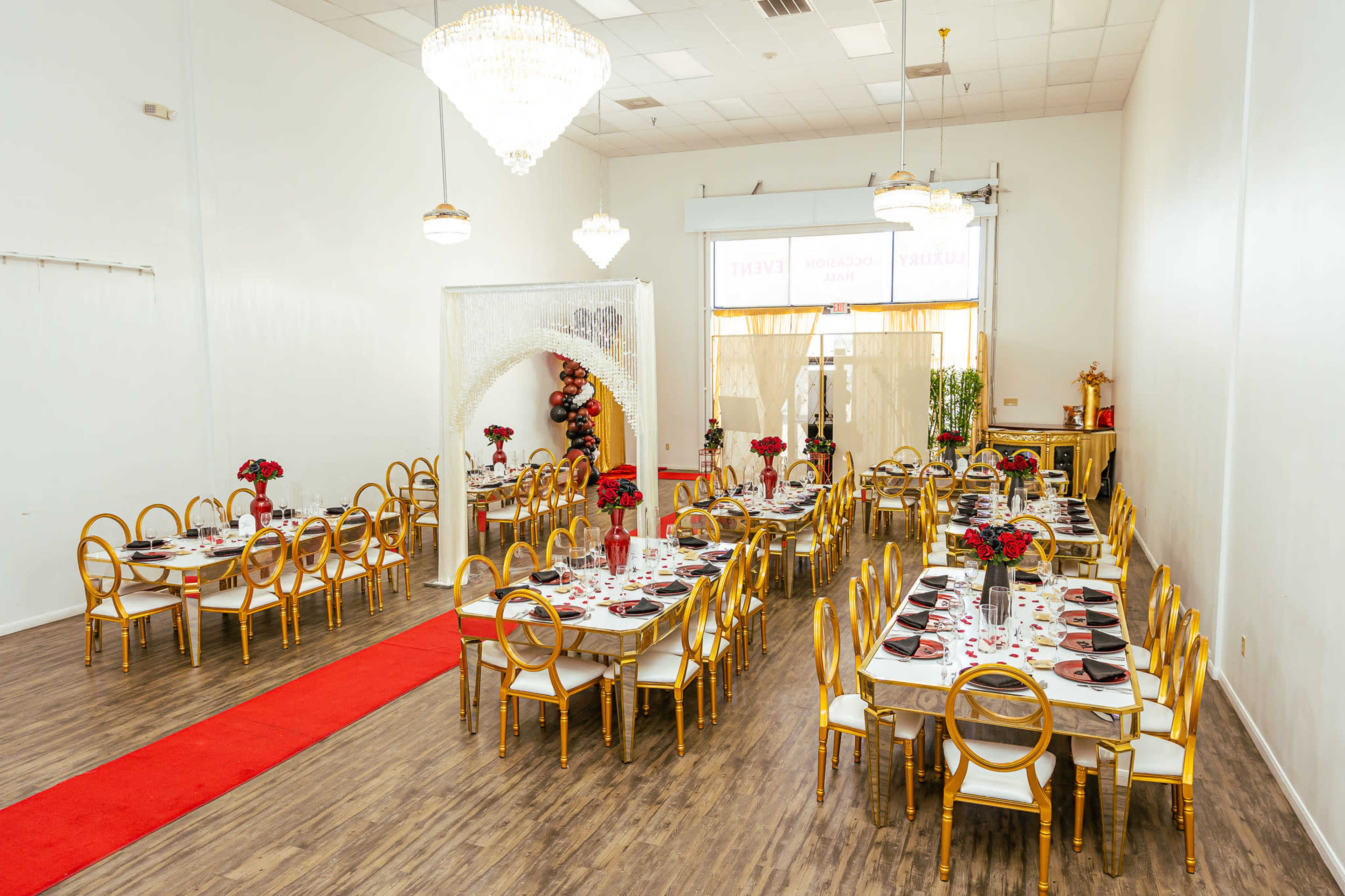 The image shows an elegantly arranged dining hall with tables set for an event, featuring gold chairs and red floral centerpieces.