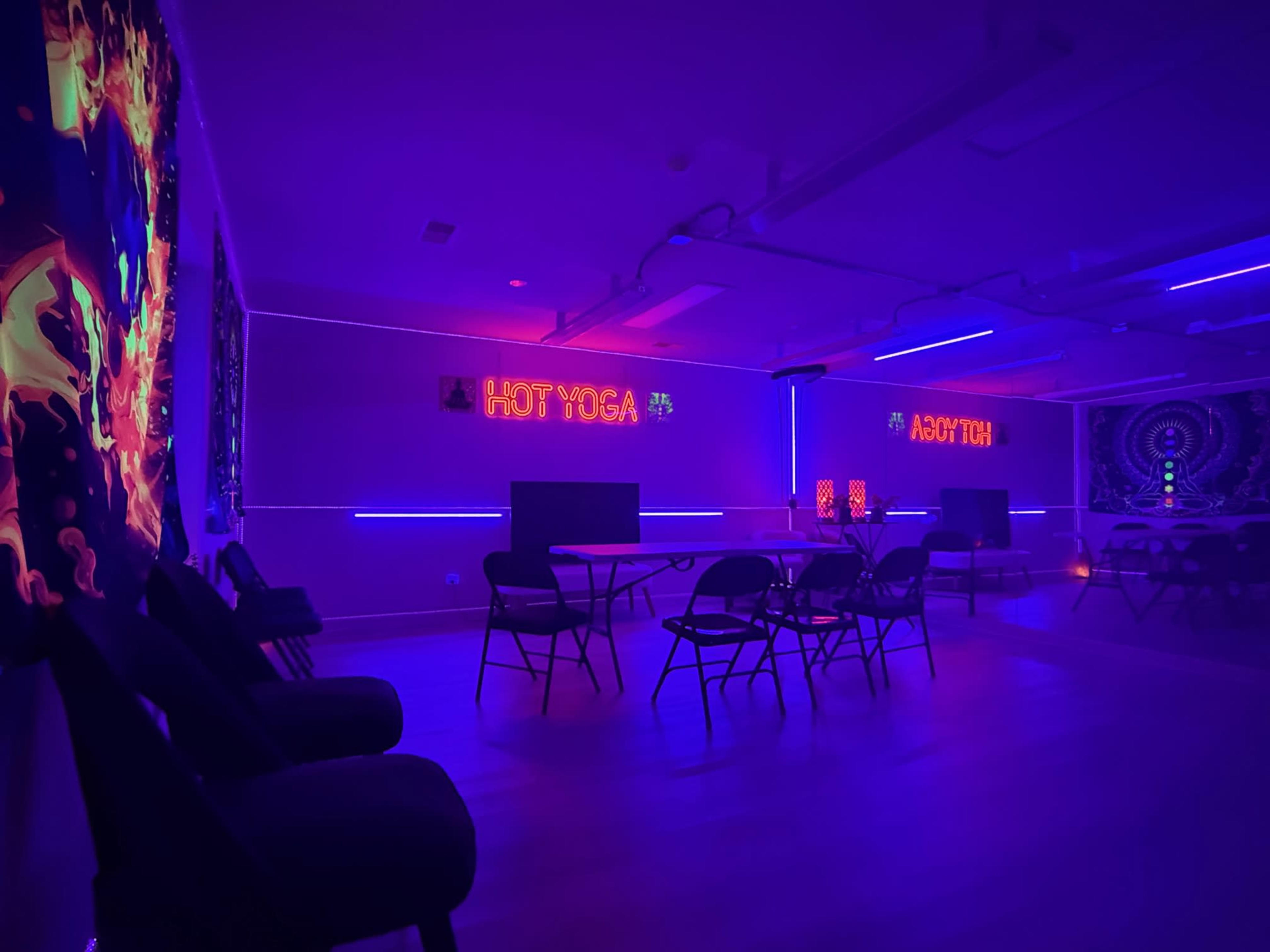 The room features neon signs and colorful lighting, with chairs arranged around tables in a yoga studio setting.