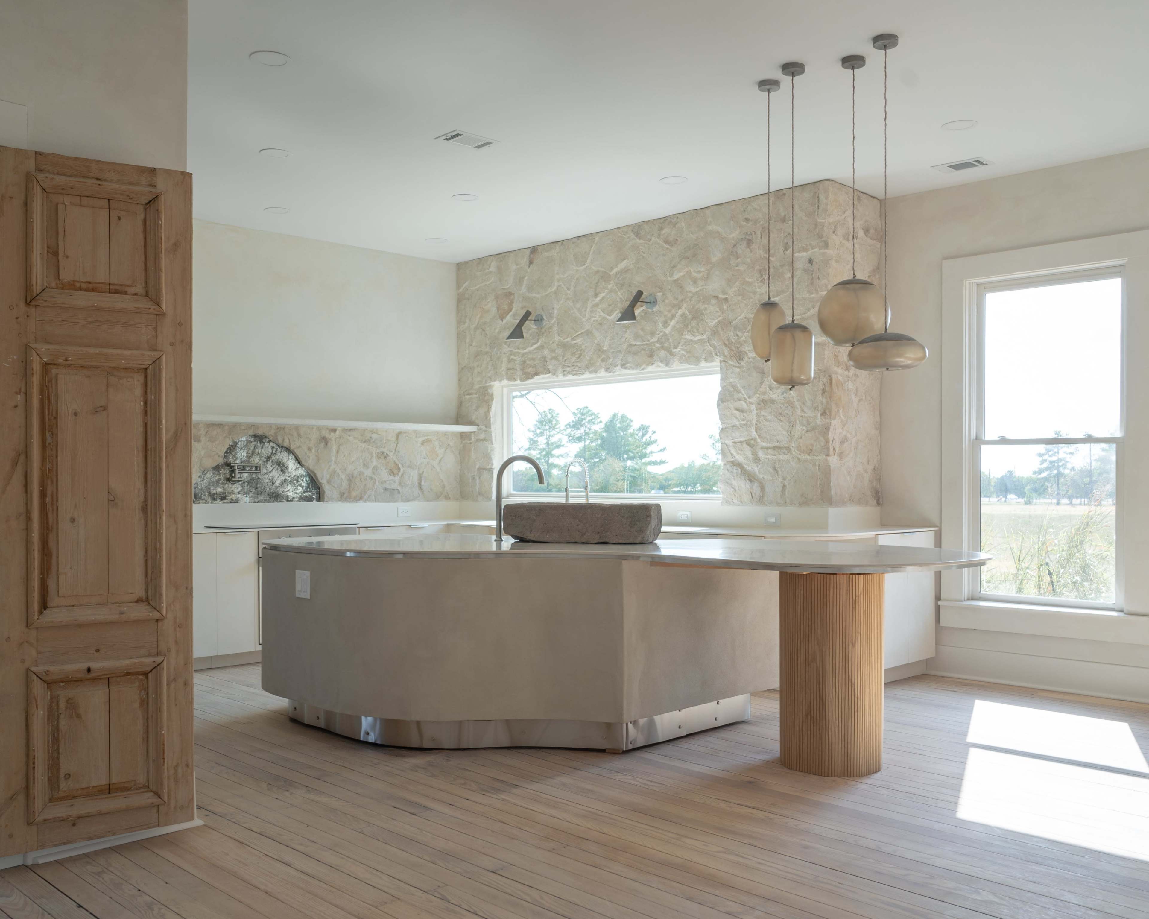 A modern kitchen with a large curved concrete island, wooden cabinetry, and stone wall accents.