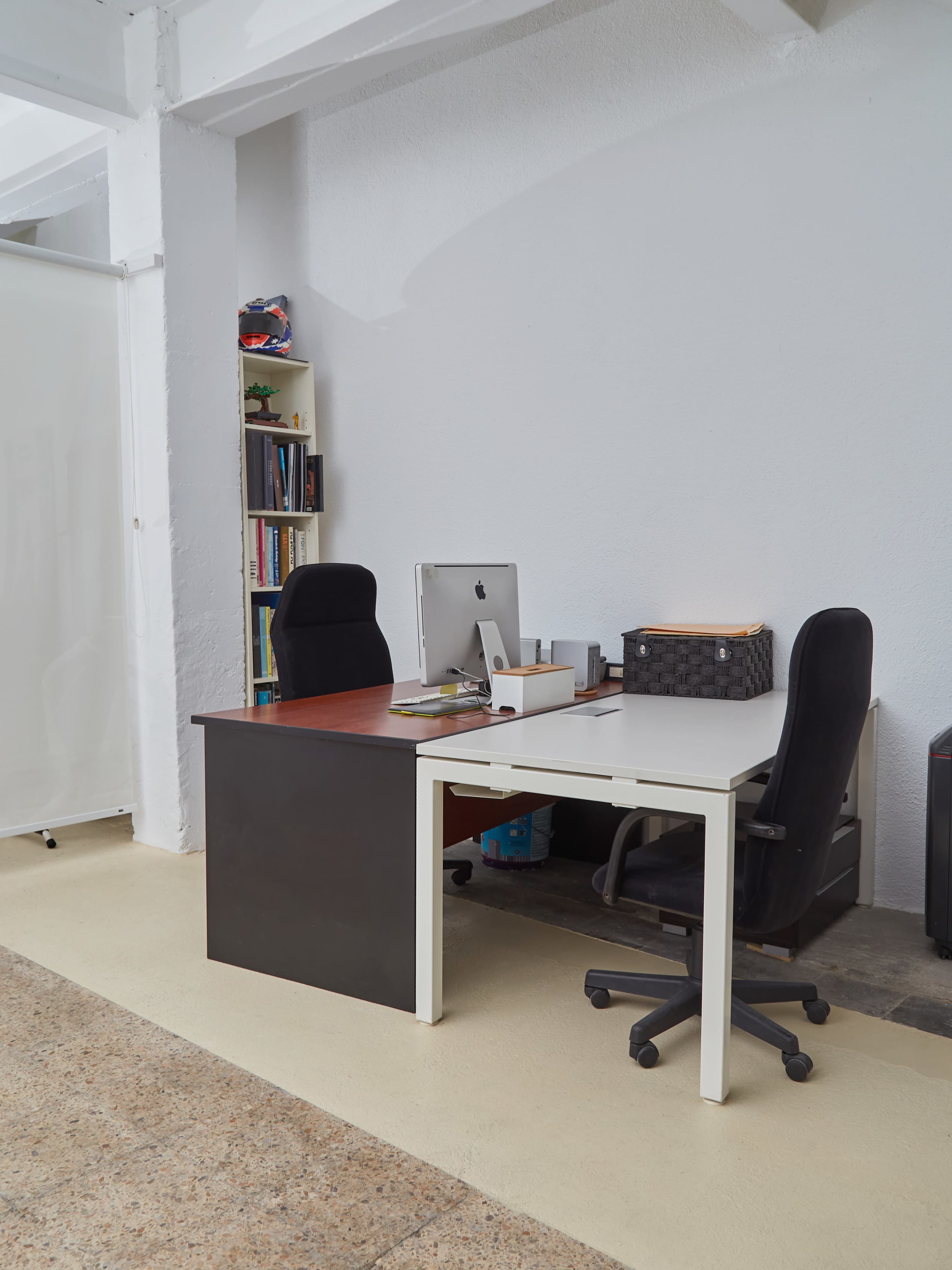 The image shows a minimalist office space with two desks, one black and one white, facing each other, accompanied by an iMac, two chairs, and a bookshelf.