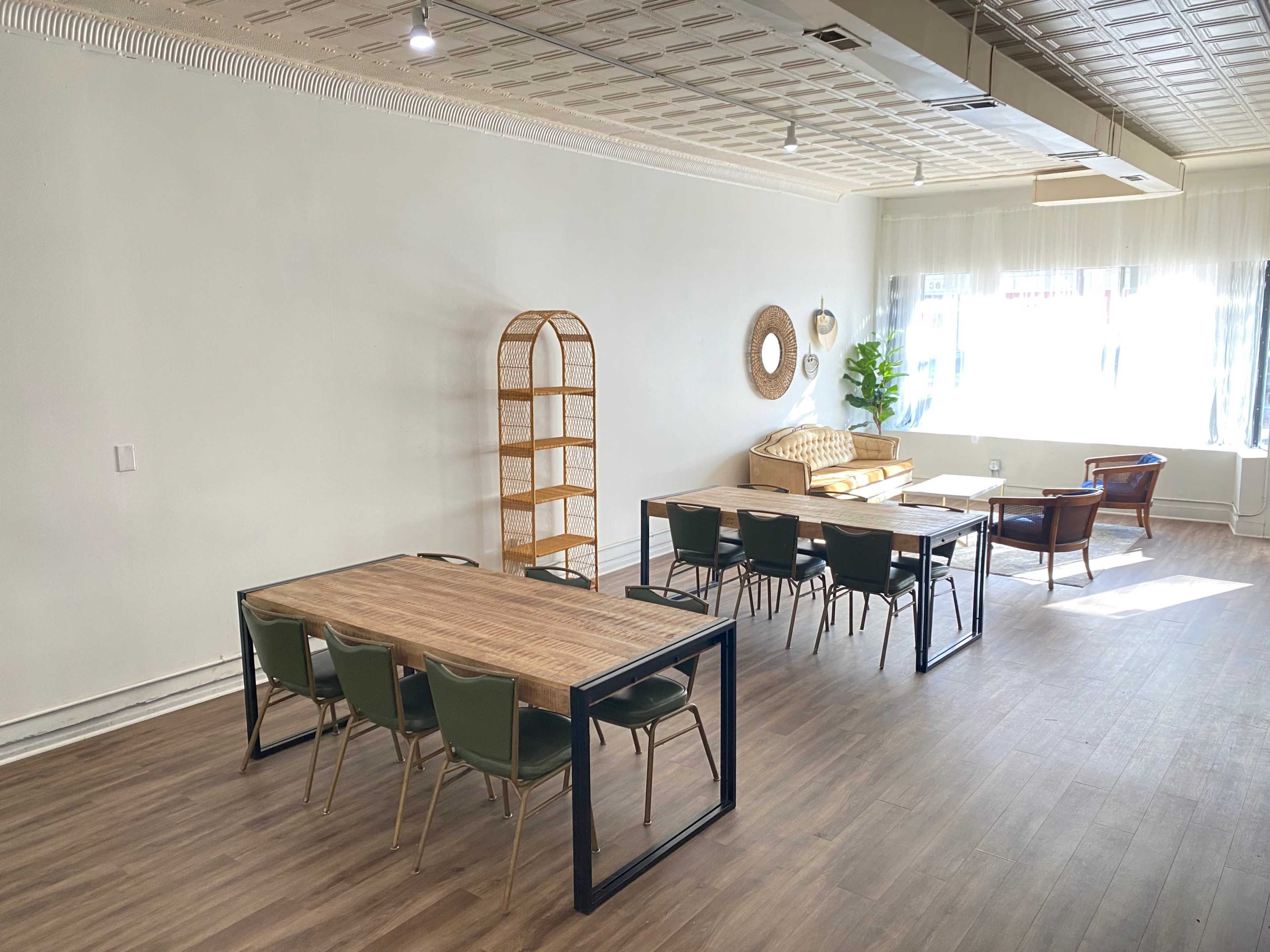 A spacious room with wooden flooring, featuring multiple tables and chairs, a wicker shelf, a sofa, and large windows allowing natural light.