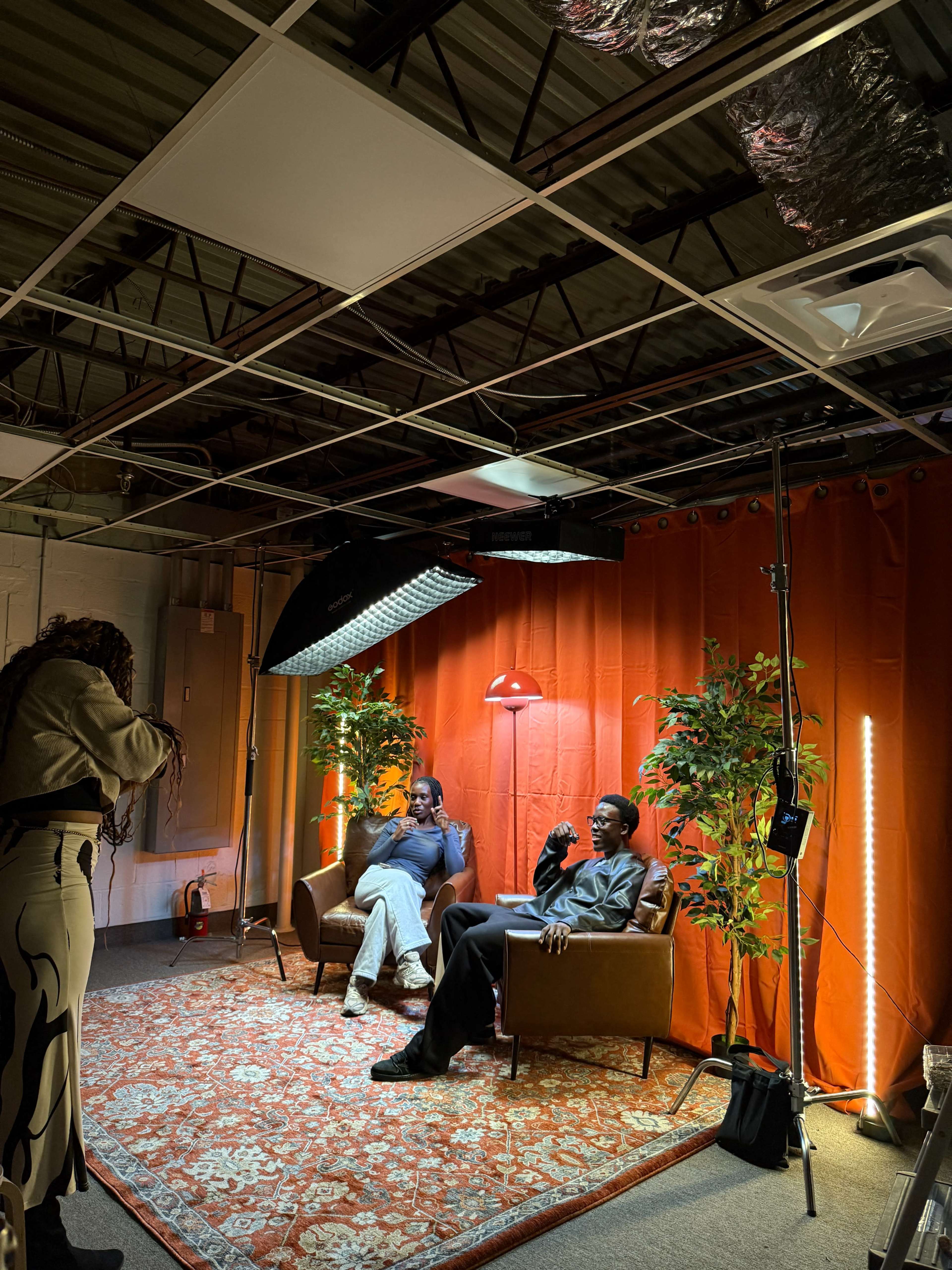 Two individuals sit in armchairs under studio lighting in a room with an orange curtain backdrop, while a third person stands nearby, facing them.