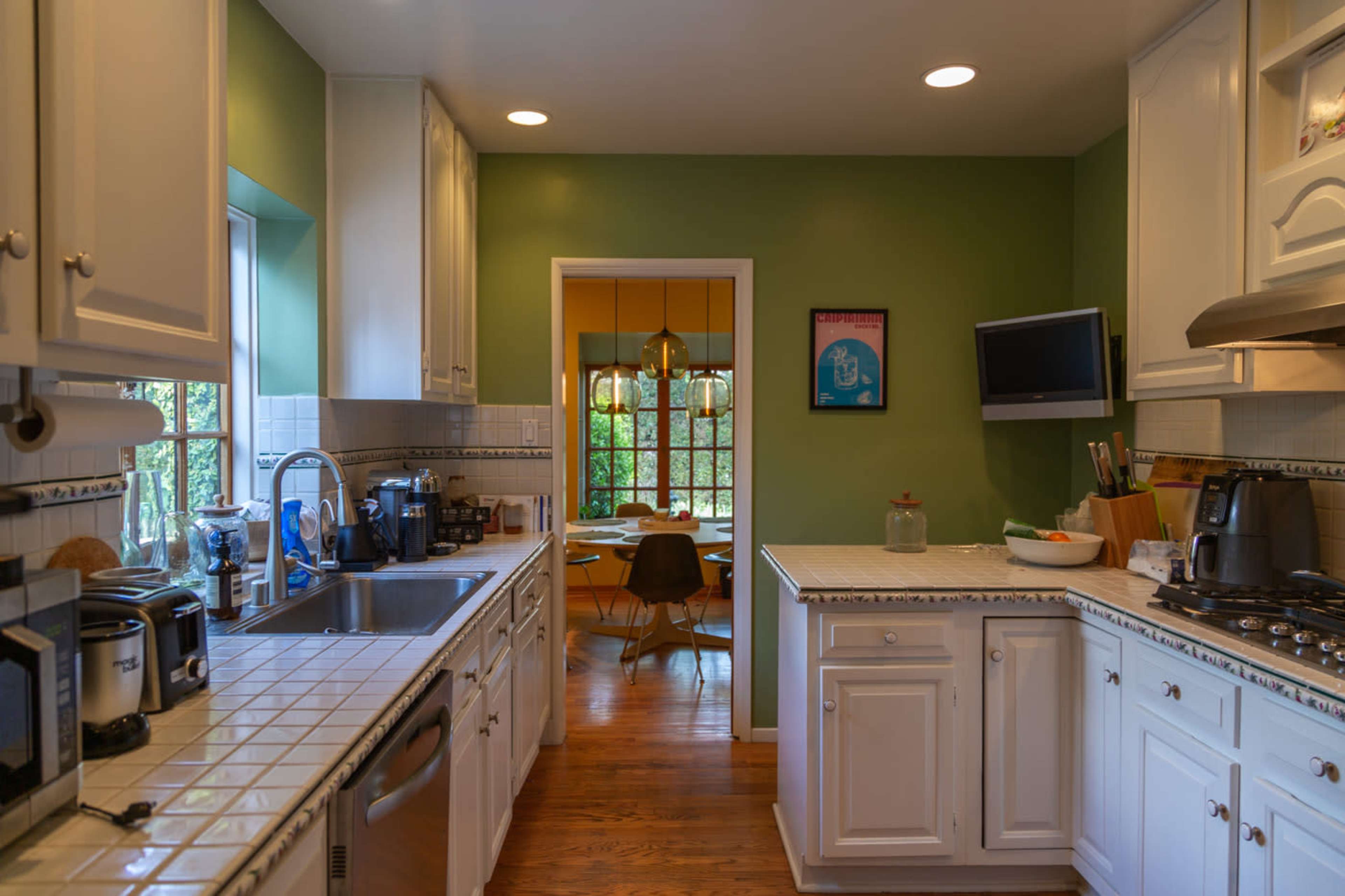 A kitchen with white cabinetry, a tiled countertop, and visible appliances leads to a dining area through a doorway, with green walls and a large window letting in natural light.