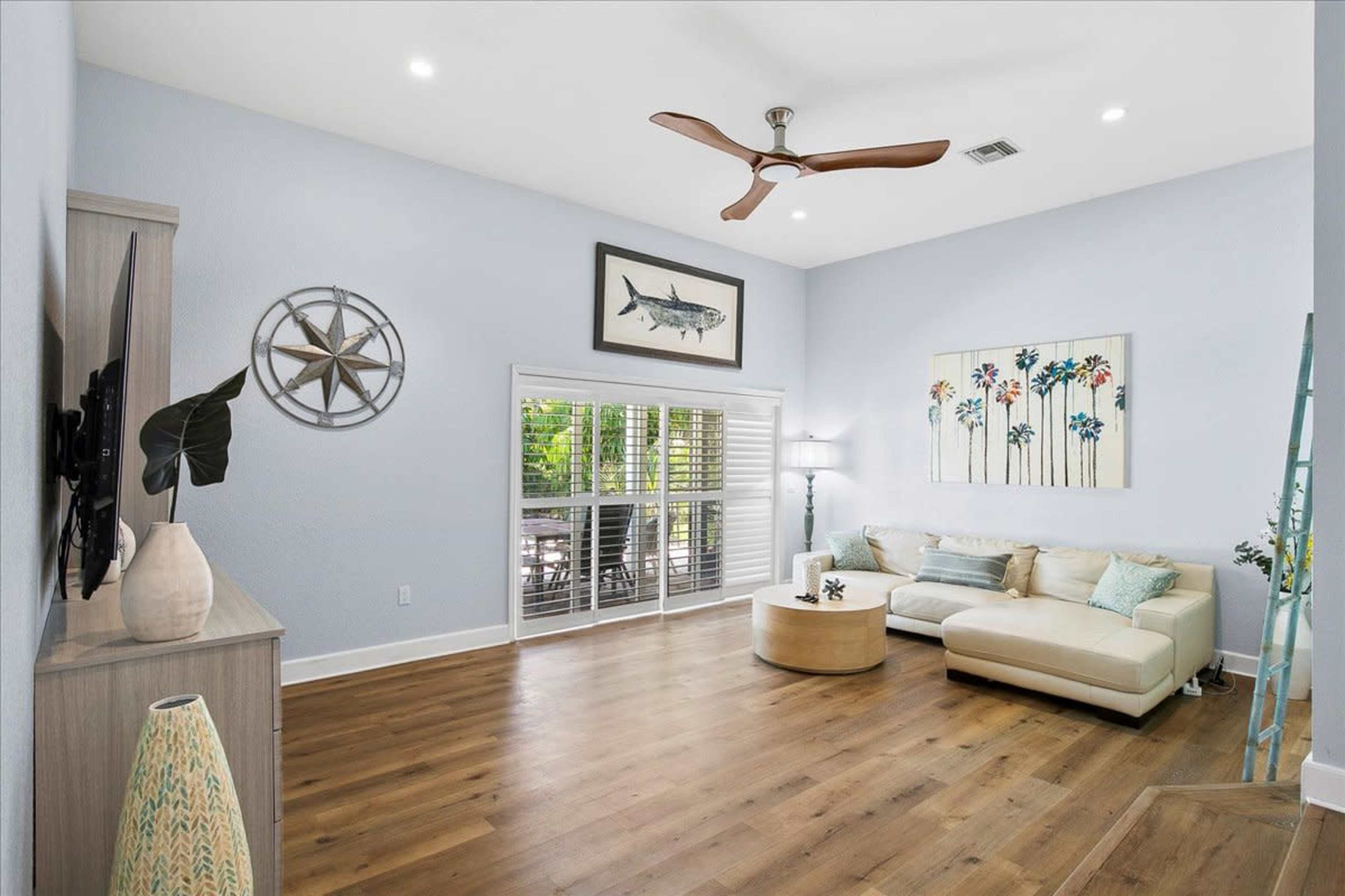 The image shows a bright living room with a light blue color scheme, featuring a beige sectional sofa, a round coffee table, and large windows adorned with plantation shutters.