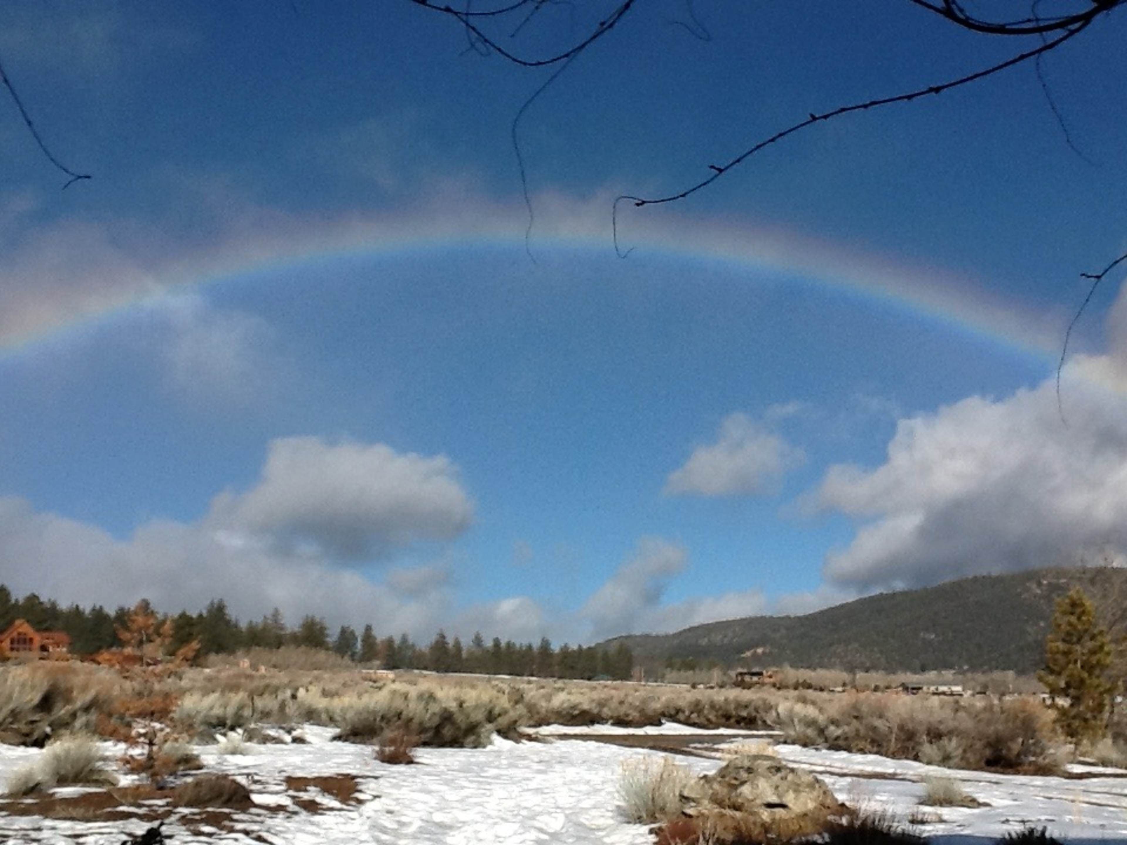A vibrant rainbow arcs over a snowy landscape with scattered trees and mountains in the background under a partly cloudy sky.