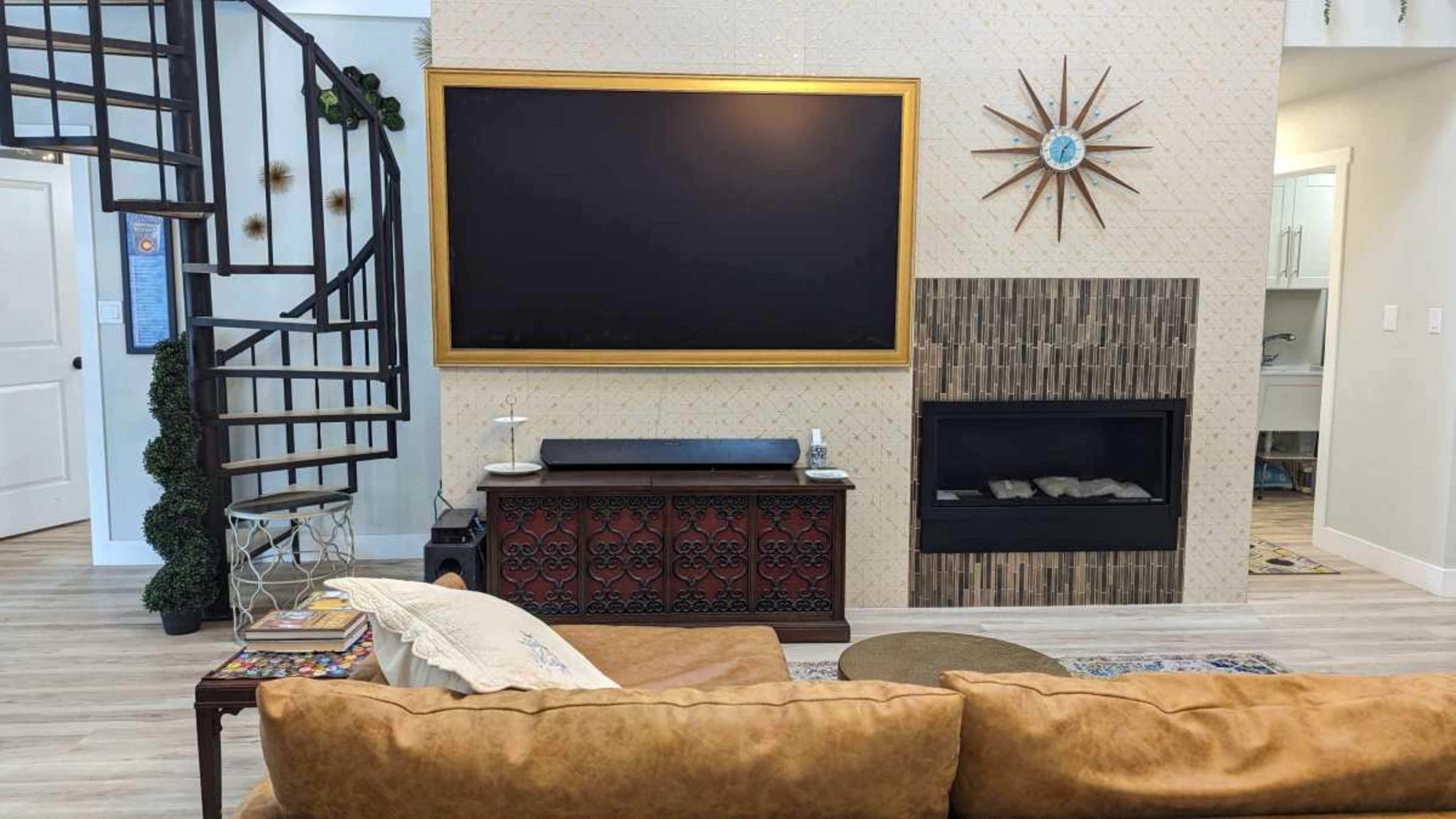 The image shows a living room with a large wall-mounted TV framed in gold, a decorative wall clock, a spiral staircase, and a modern fireplace below a TV unit.