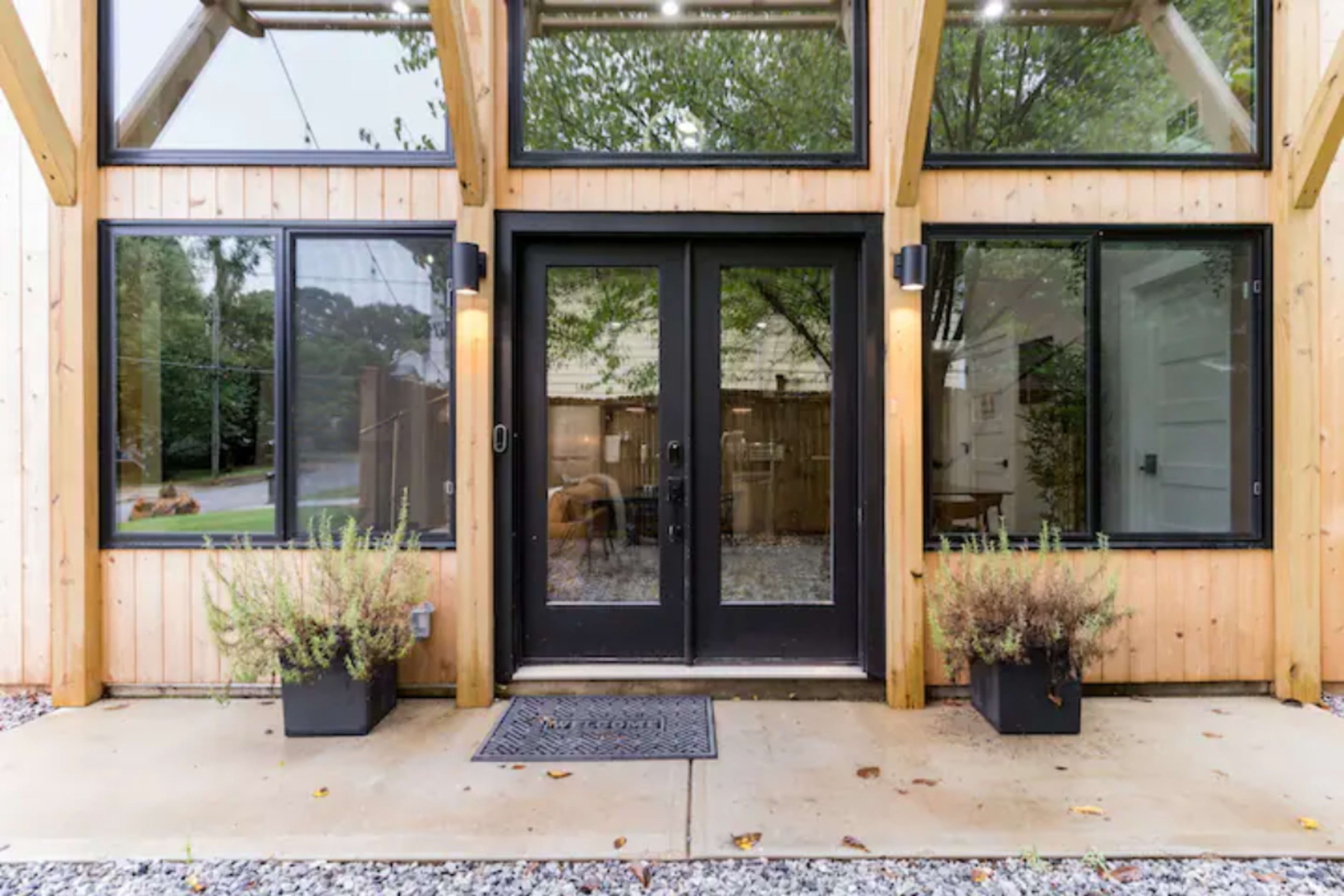The image shows a modern wooden structure with large glass doors flanked by potted plants and gravel landscaping.