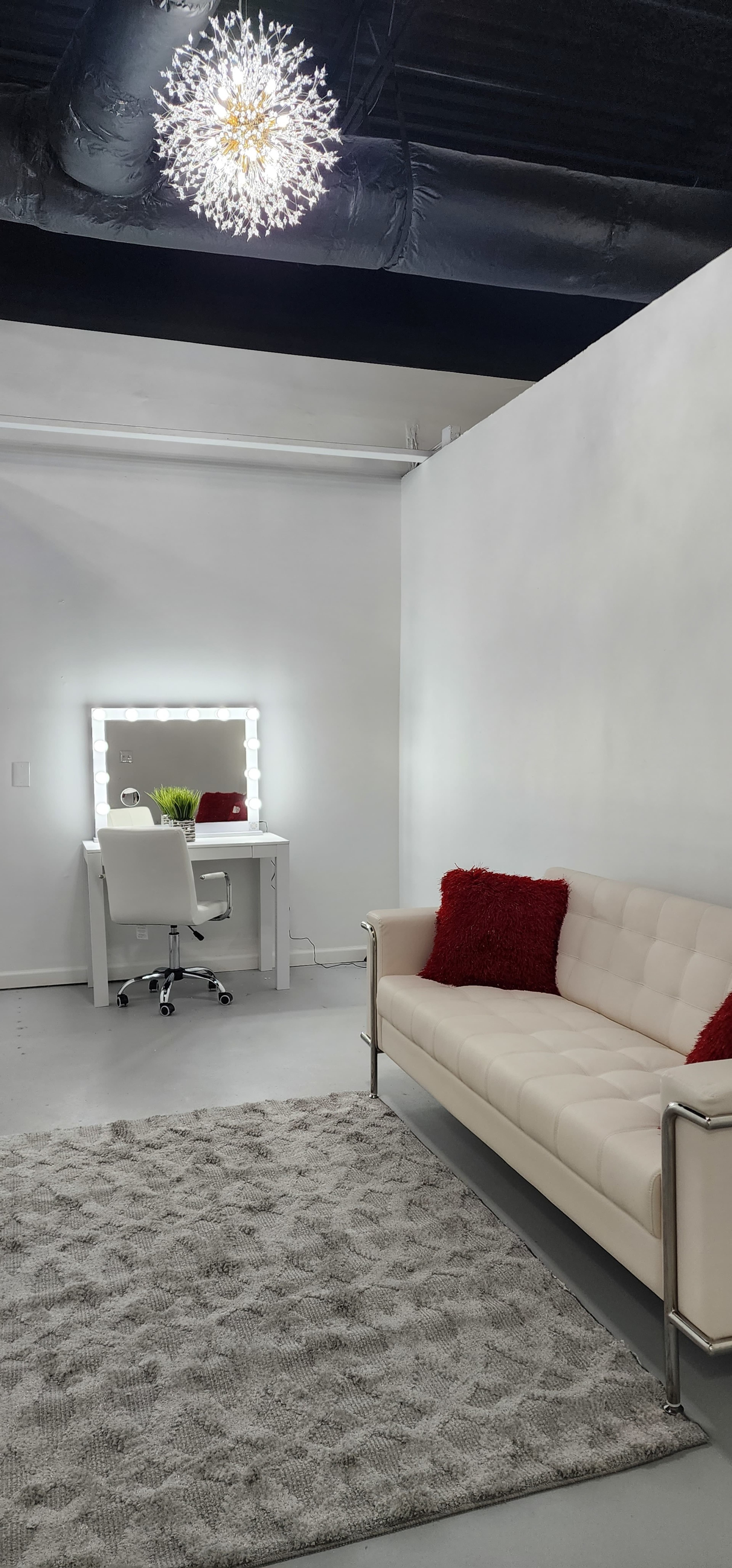 The image shows a modern, minimalist interior featuring a white couch, a large rug, a vanity with a mirror and lights, and a small plant on a desk.