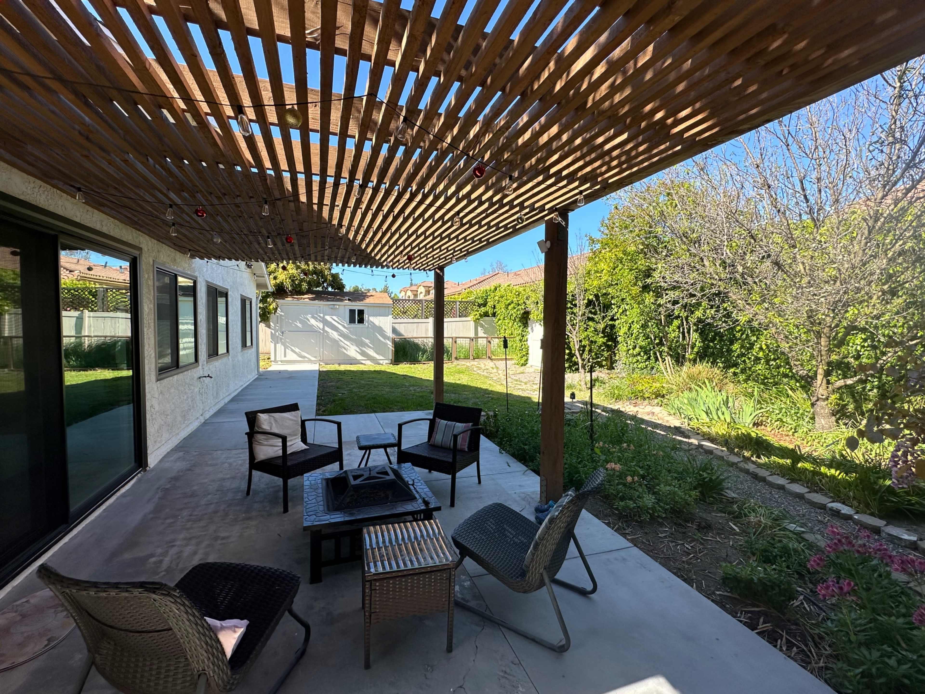 A patio area with seating furniture arranged around a central fire pit, overlooking a landscaped garden with grass and plants.