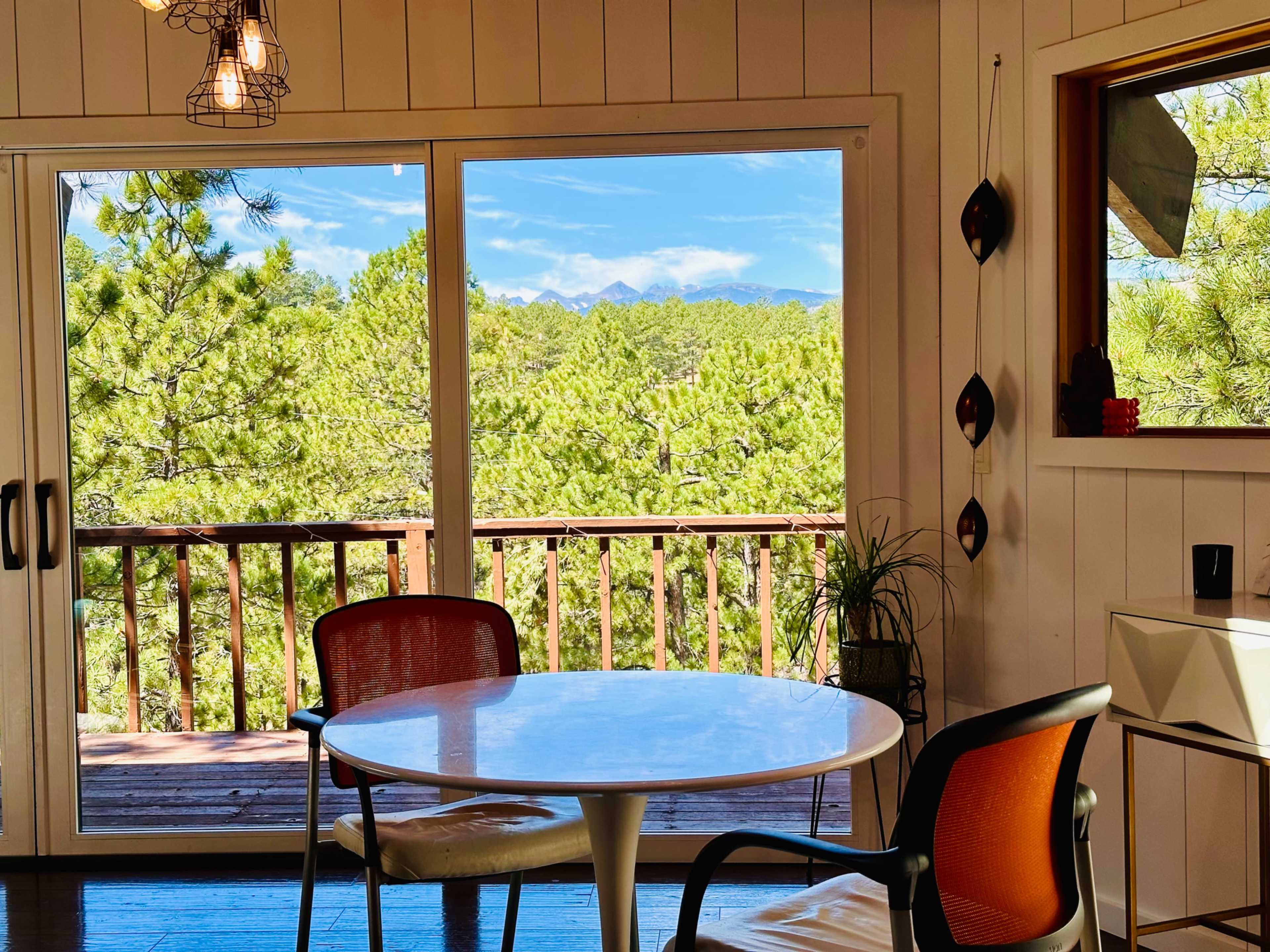 A small dining area features a round table with two chairs, surrounded by large windows that offer a view of pine trees and distant mountains.