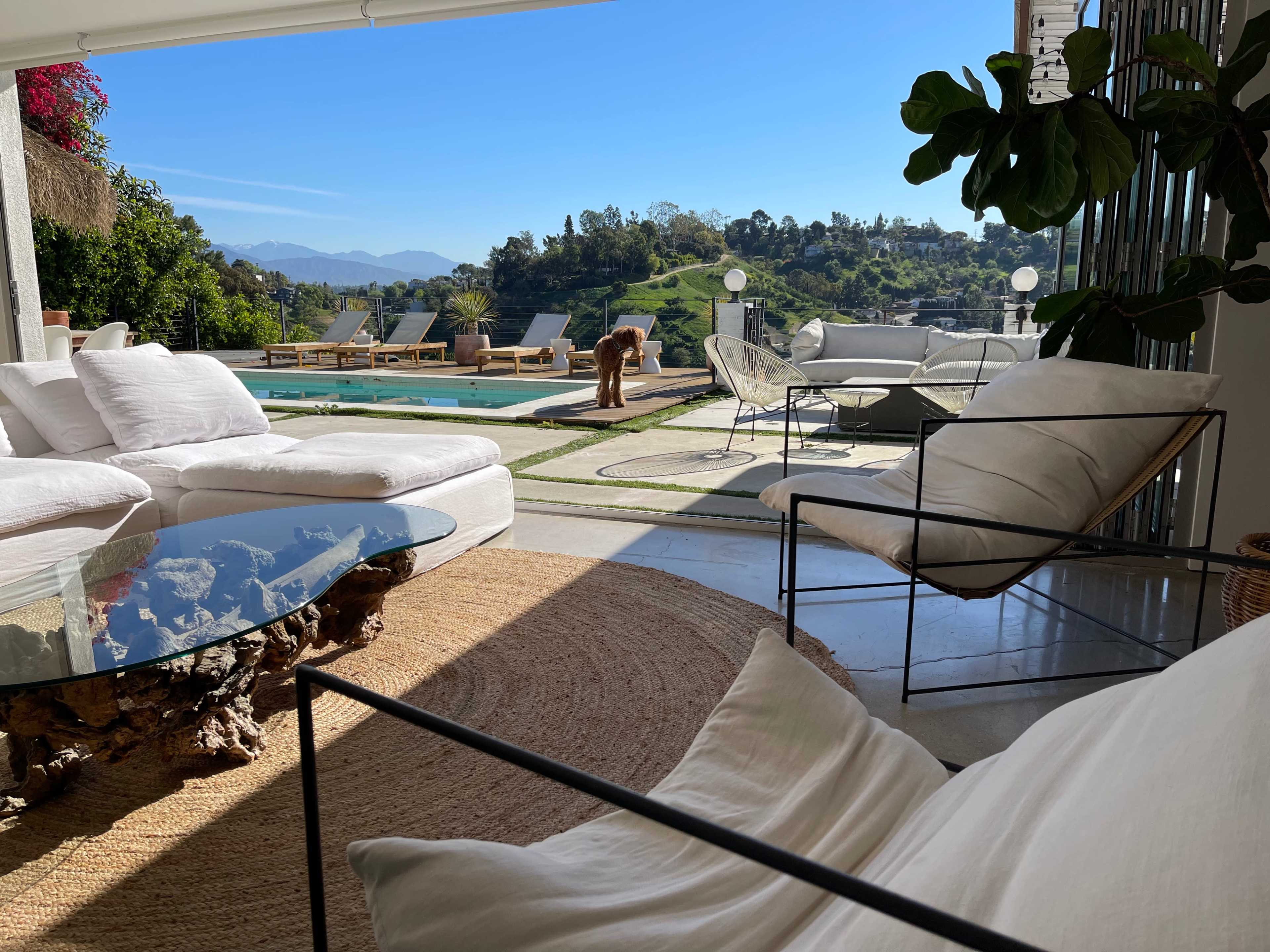 A sunlit patio features modern furniture overlooking a swimming pool and green hills in the background.