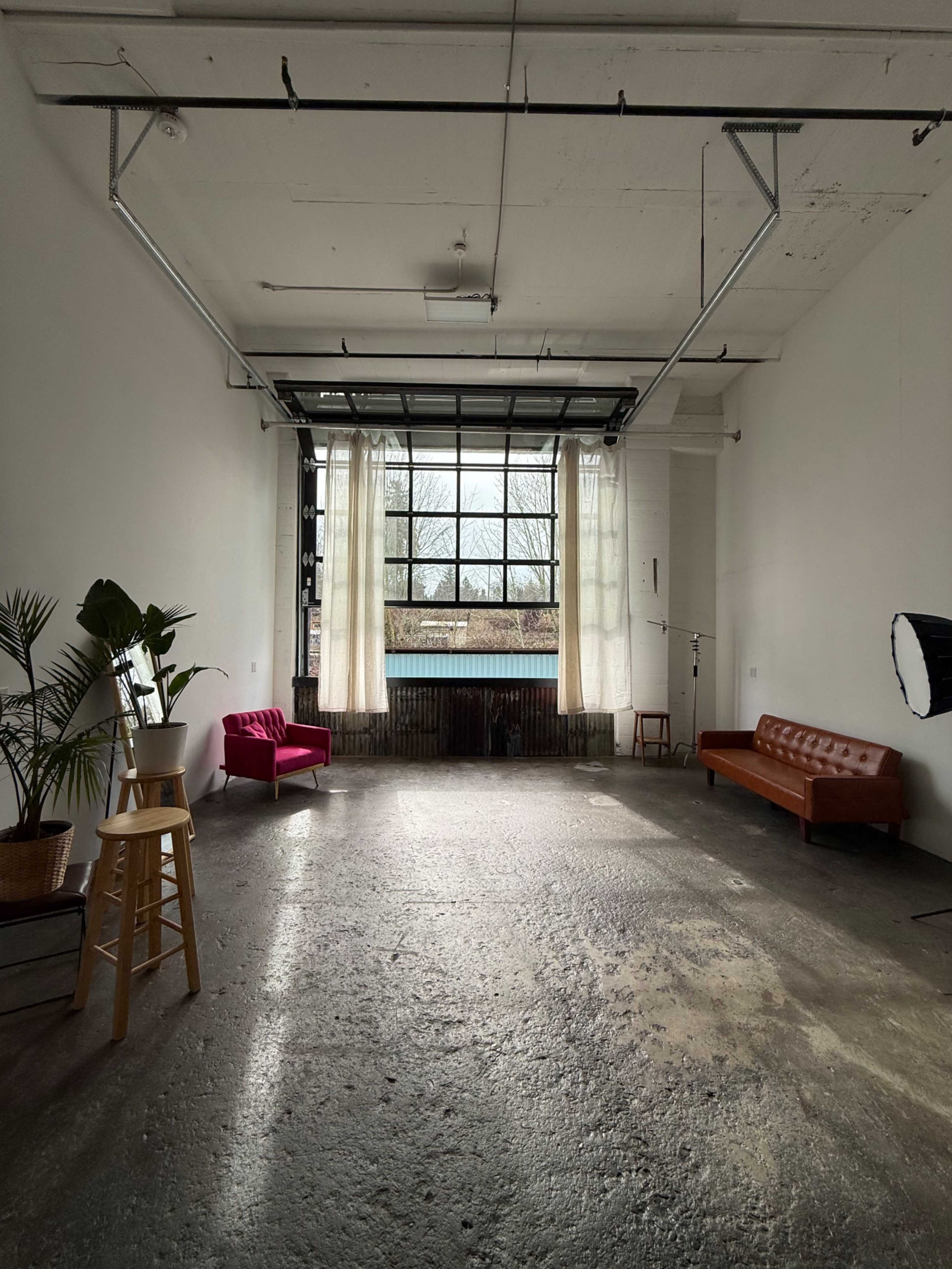 The image shows a spacious, empty studio with large windows, a brown leather couch, a pink chair, a plant, and a light stand.