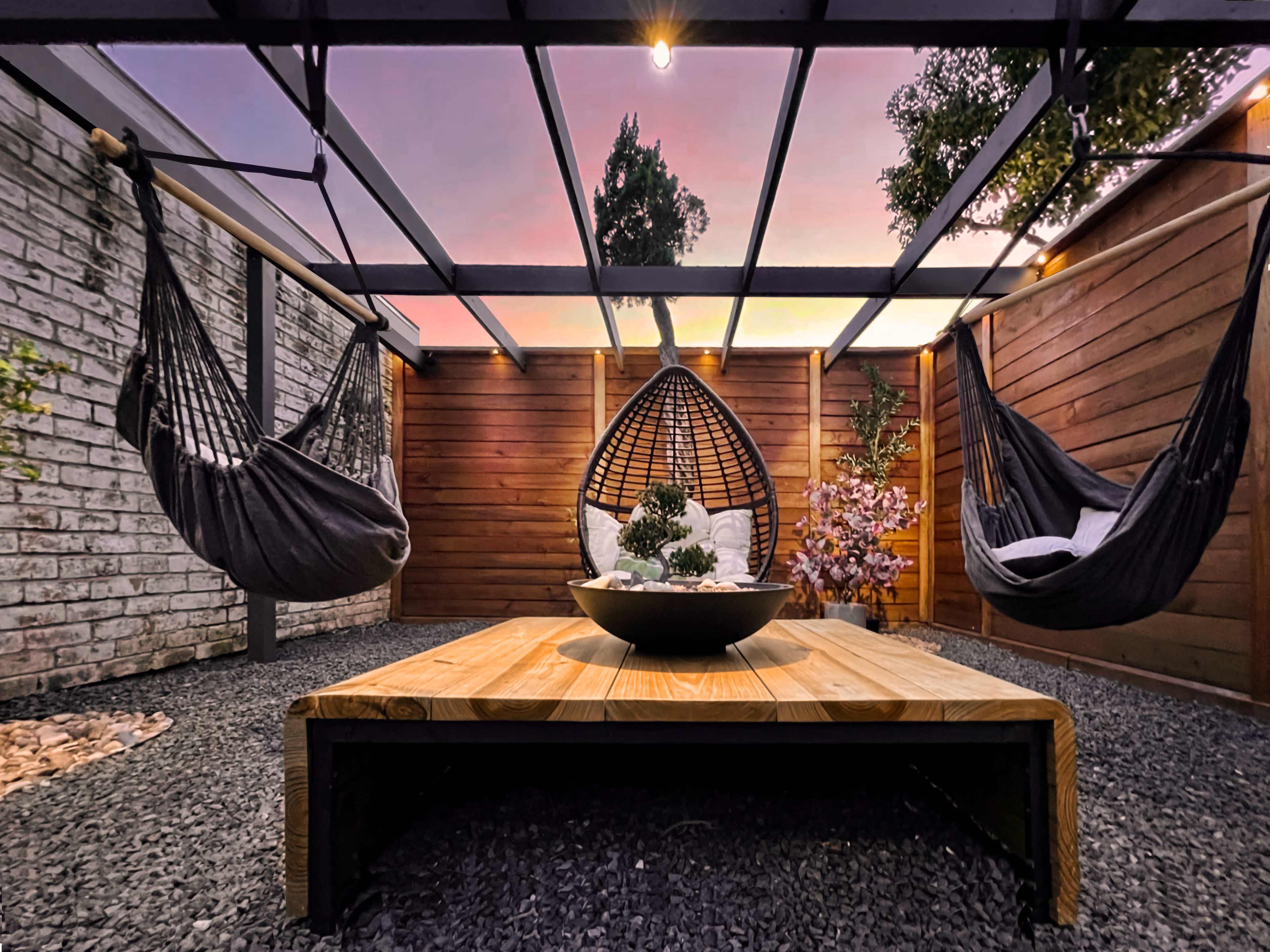 A modern outdoor space features three hanging chairs, a wooden table, and decorative plants under a wooden pergola at sunset.