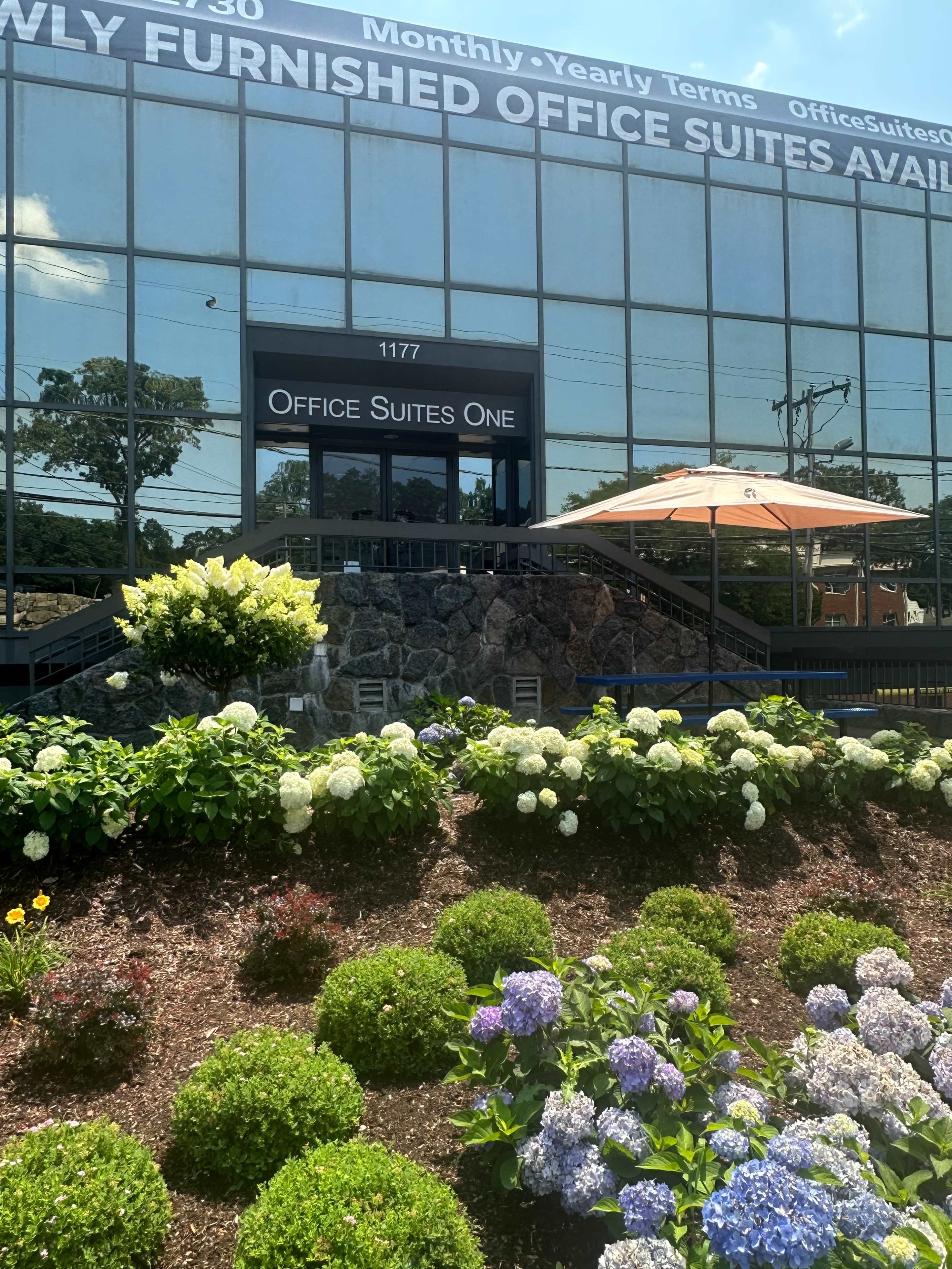A modern office building with large glass windows, featuring a sign that reads "Office Suites One" and surrounded by well-maintained green landscaping and colorful flowers.