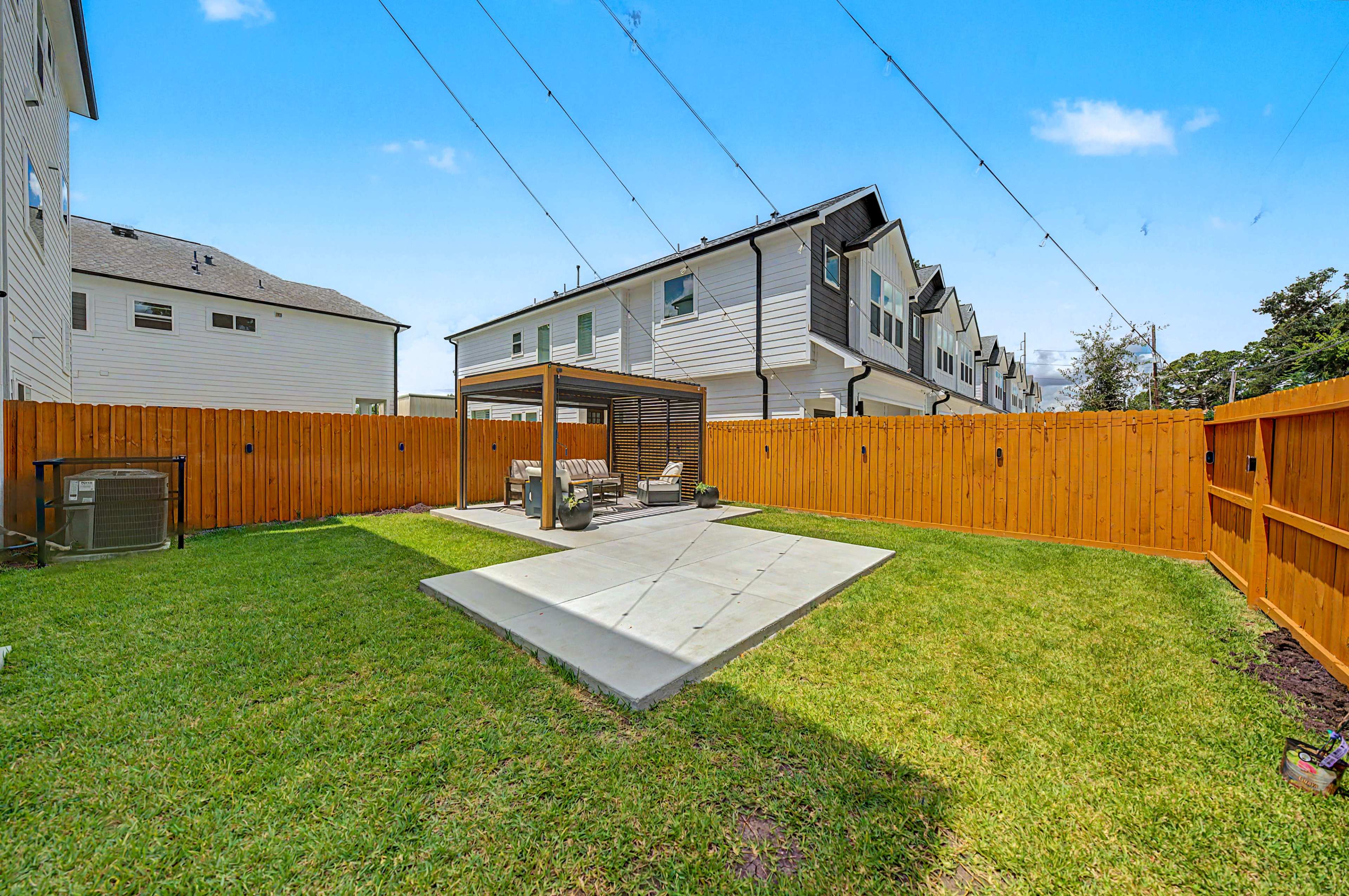 The image shows a fenced backyard featuring a concrete patio with outdoor furniture and a pergola, surrounded by green grass and residential buildings.