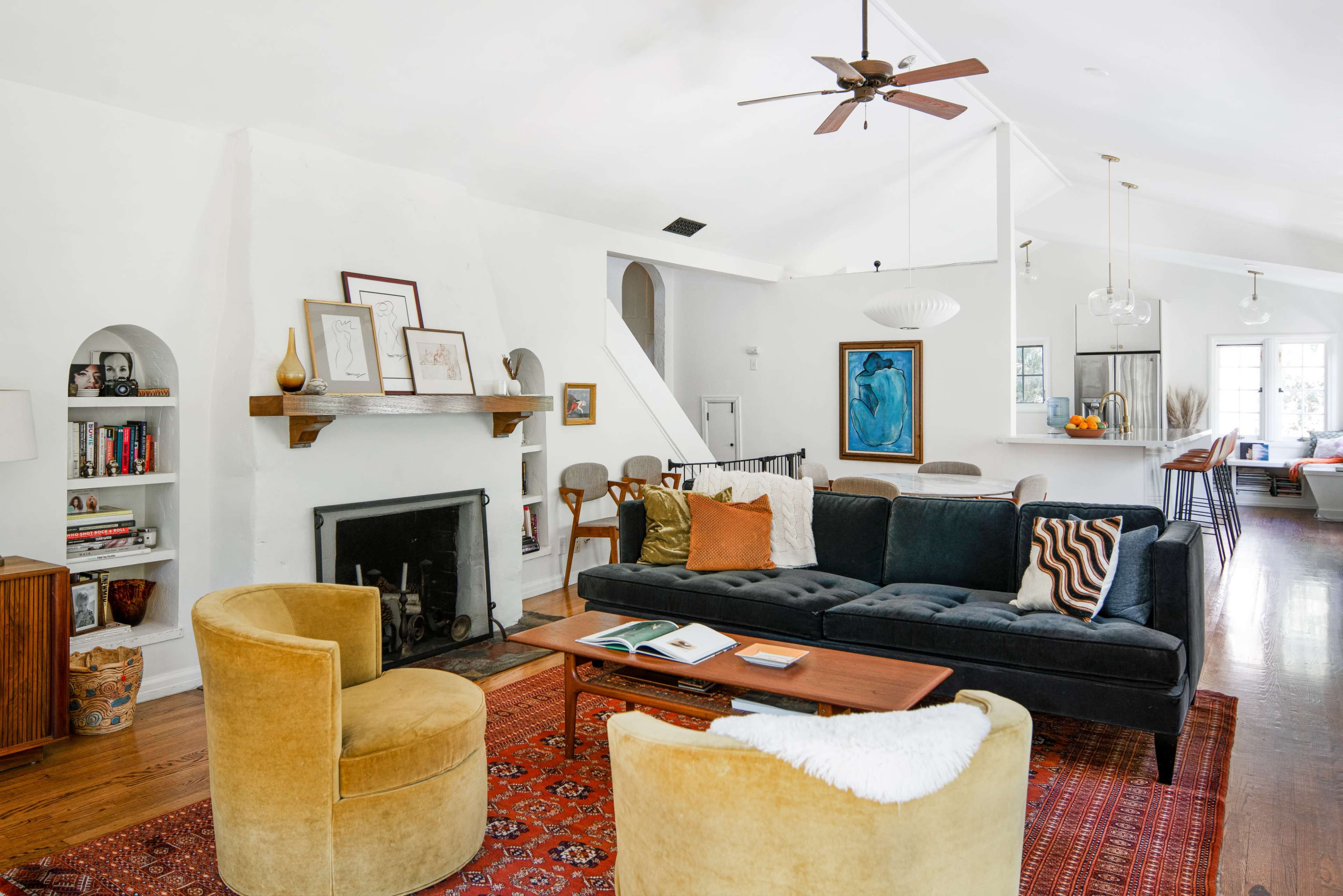 The image shows a modern living room with a dark couch, two yellow armchairs, a wooden coffee table, and a fireplace, featuring white walls and wooden floors.