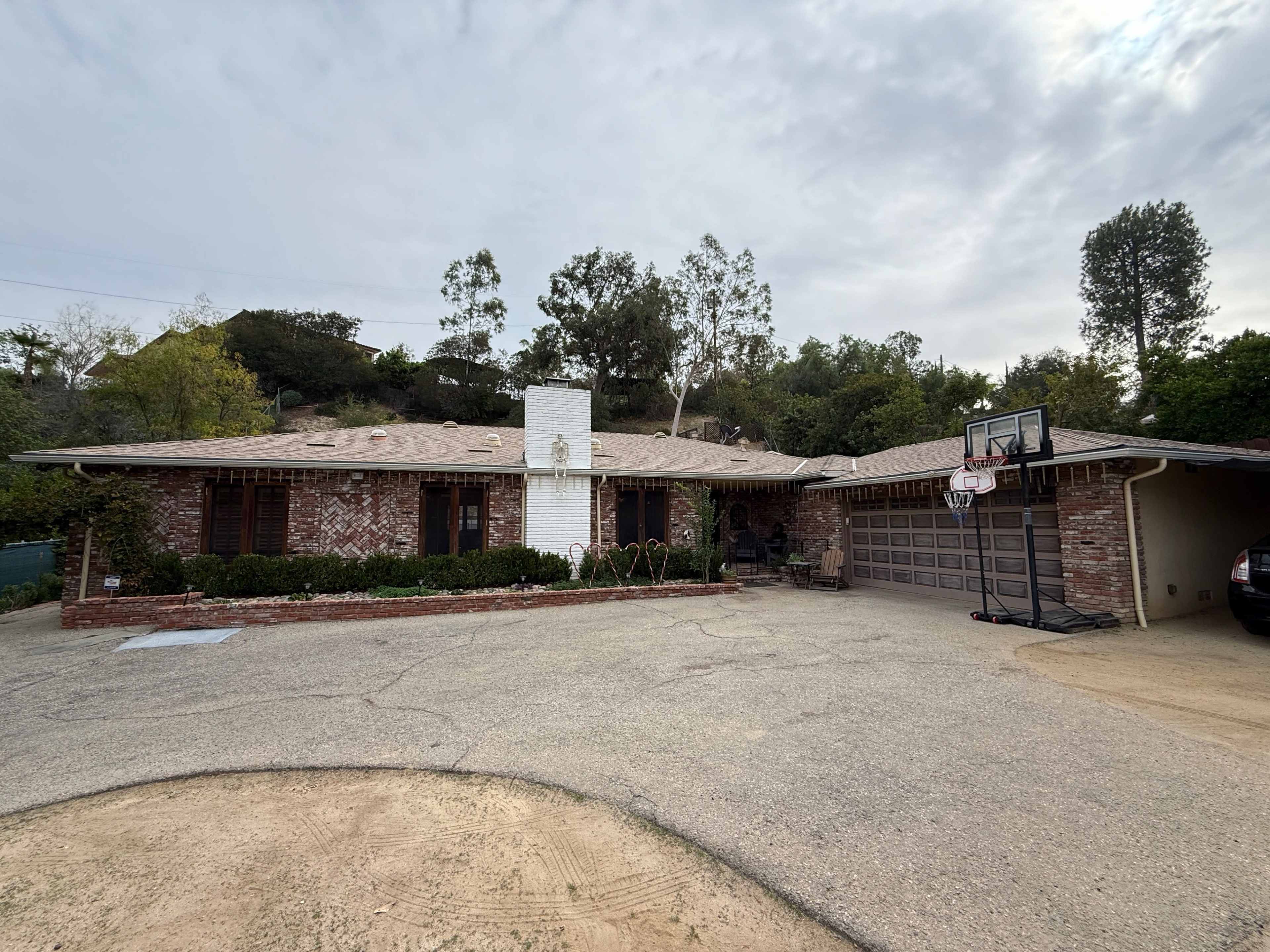 The image shows a single-story brick house with a basketball hoop in the driveway, surrounded by greenery and a gravel pathway.