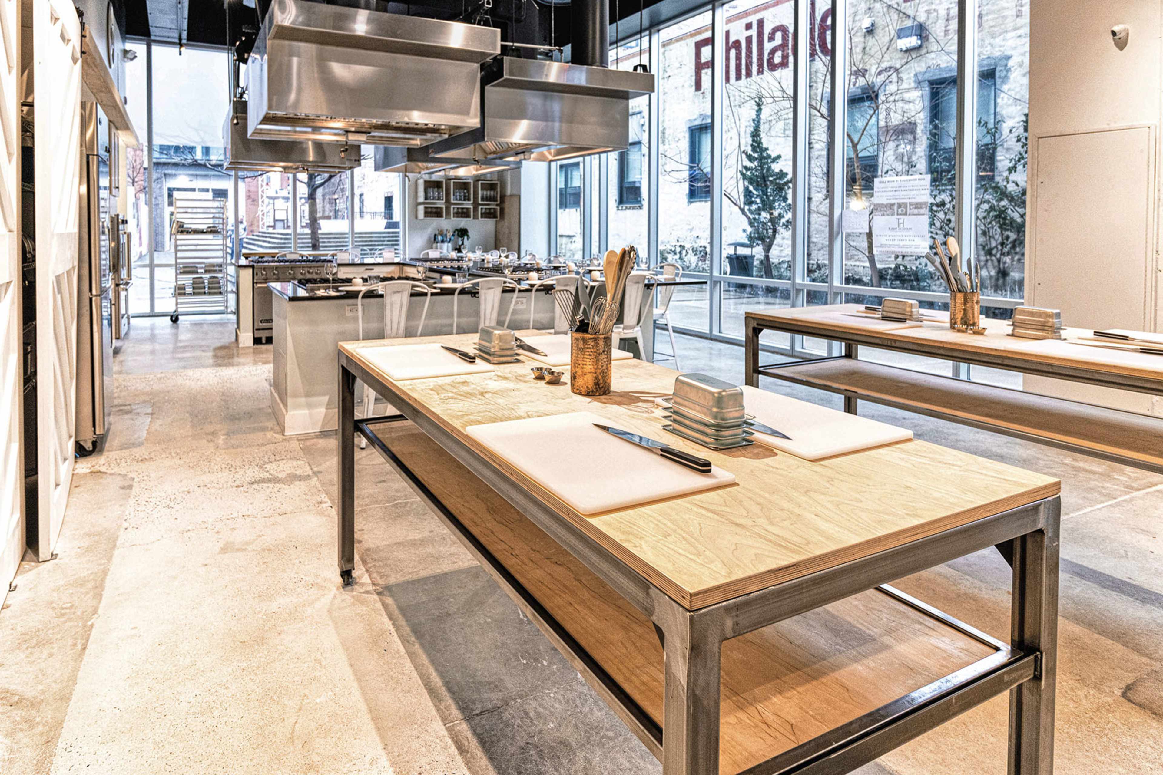 A spacious kitchen workspace with large wooden tables, stainless steel appliances, and natural light filtering through large windows.