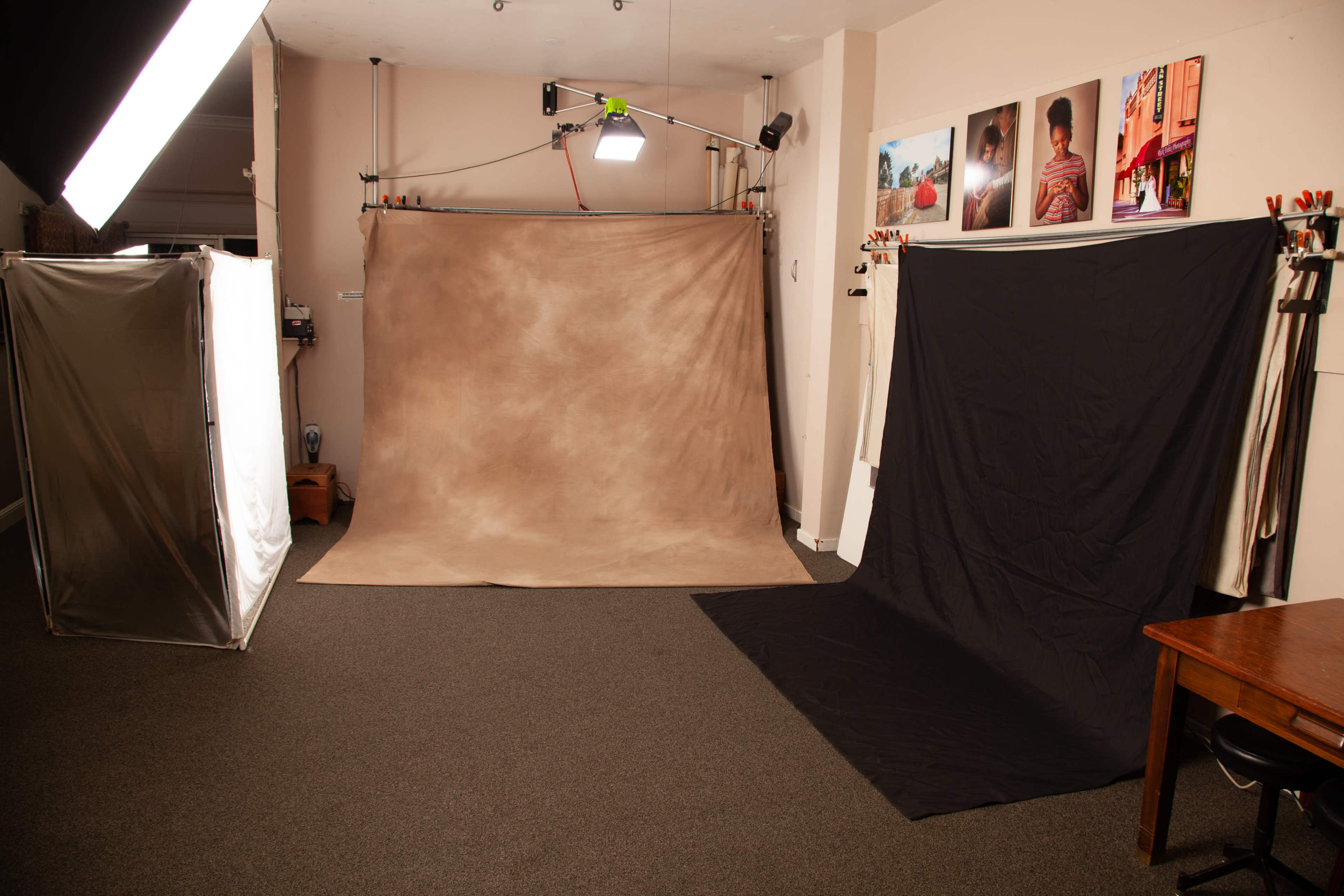 The image shows a photography studio with a backdrop setup, including a brown and a black backdrop, along with lighting equipment and photographs displayed on the wall.