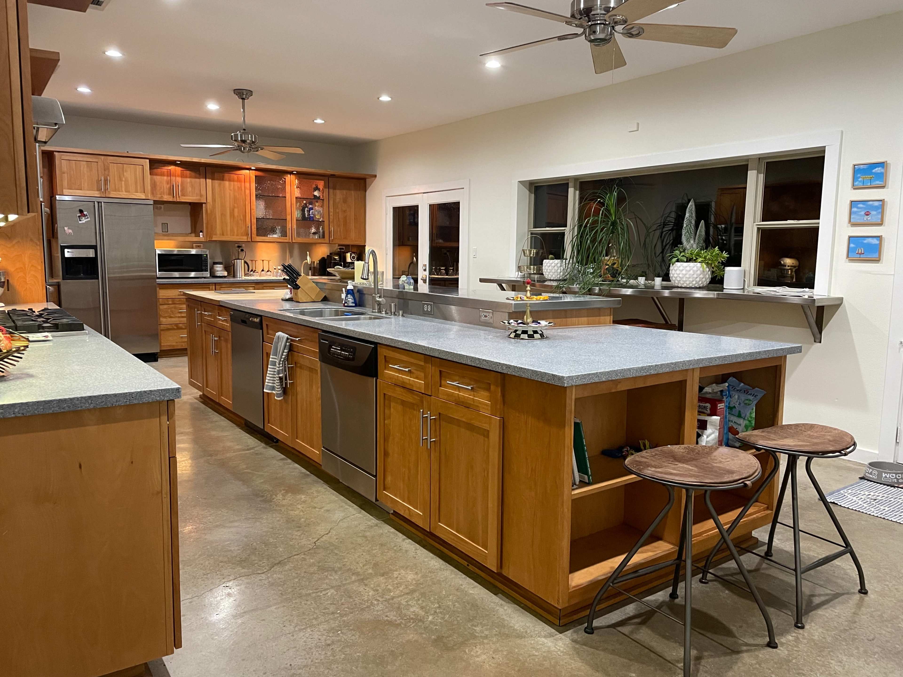 A modern kitchen features wooden cabinetry, a large central island with a gray countertop, stainless steel appliances, and two stools.