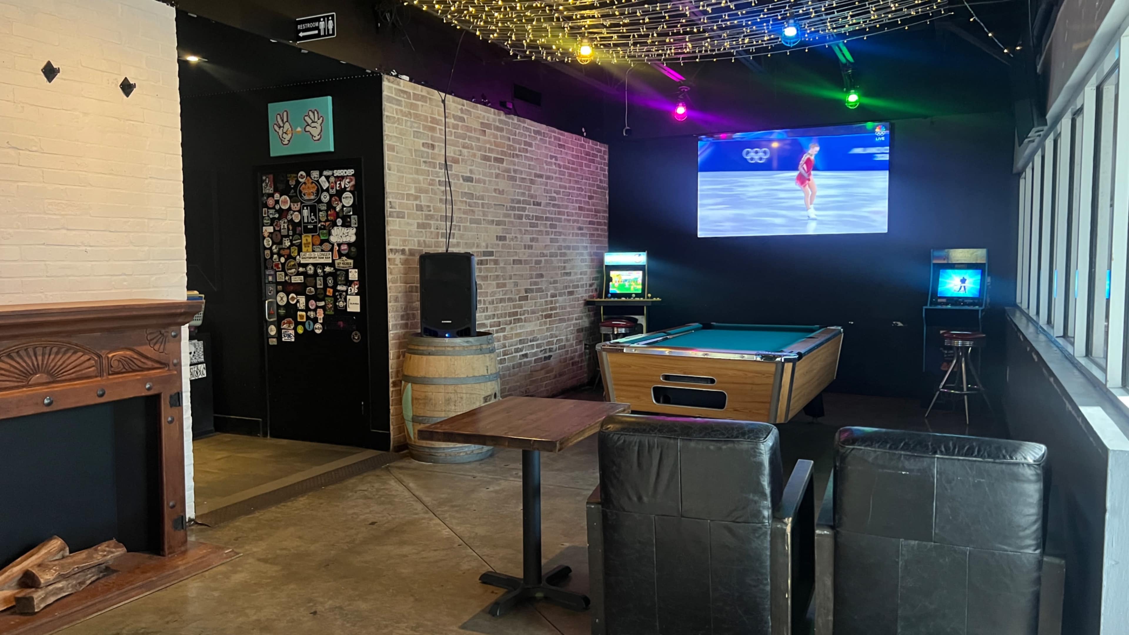 The image shows a lounge area with two leather chairs, a pool table, and a screen displaying an Olympic event, all set against a brick wall adorned with colorful lights.