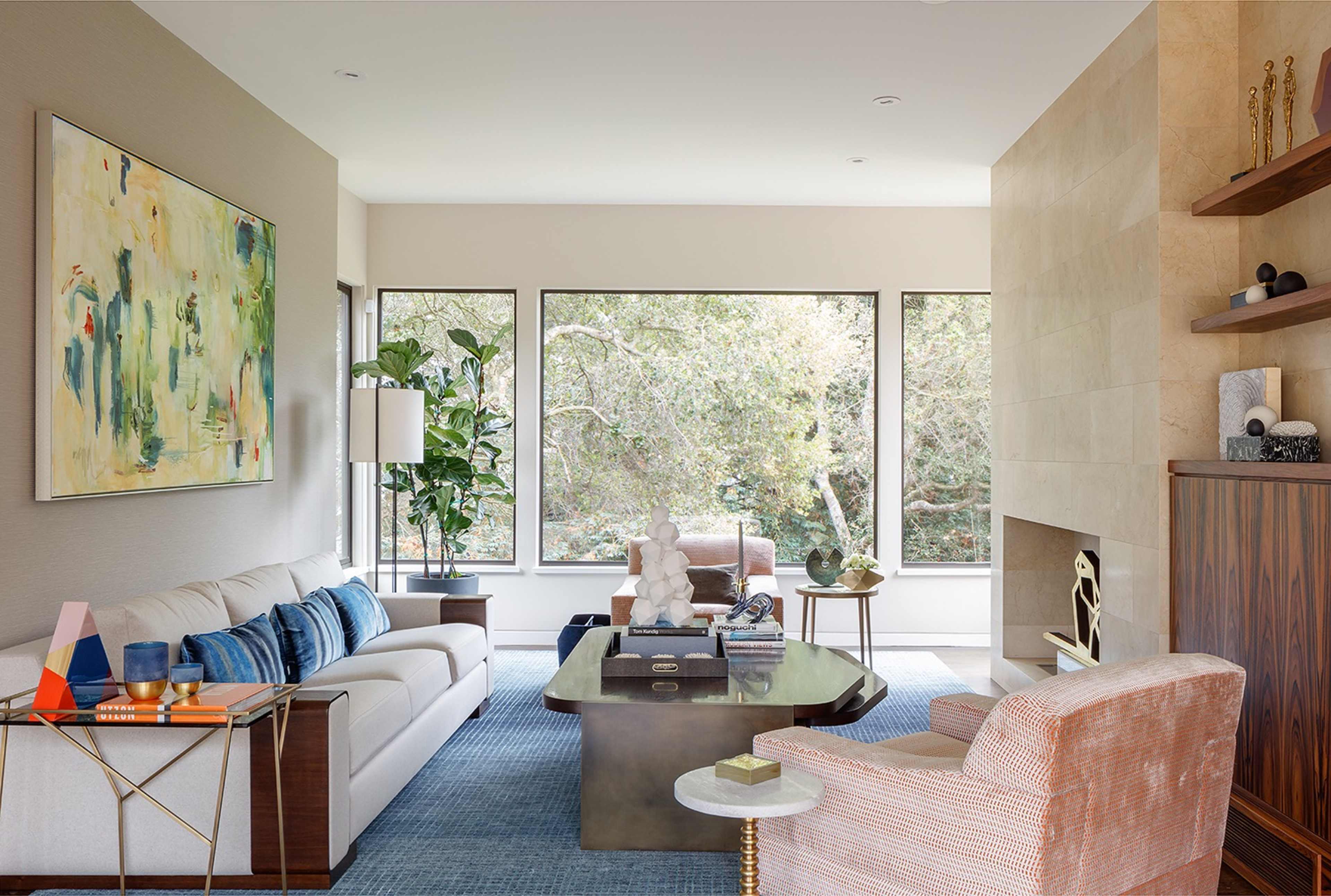 A modern living room features a white sofa, a coffee table, a teal rug, and large windows overlooking greenery.