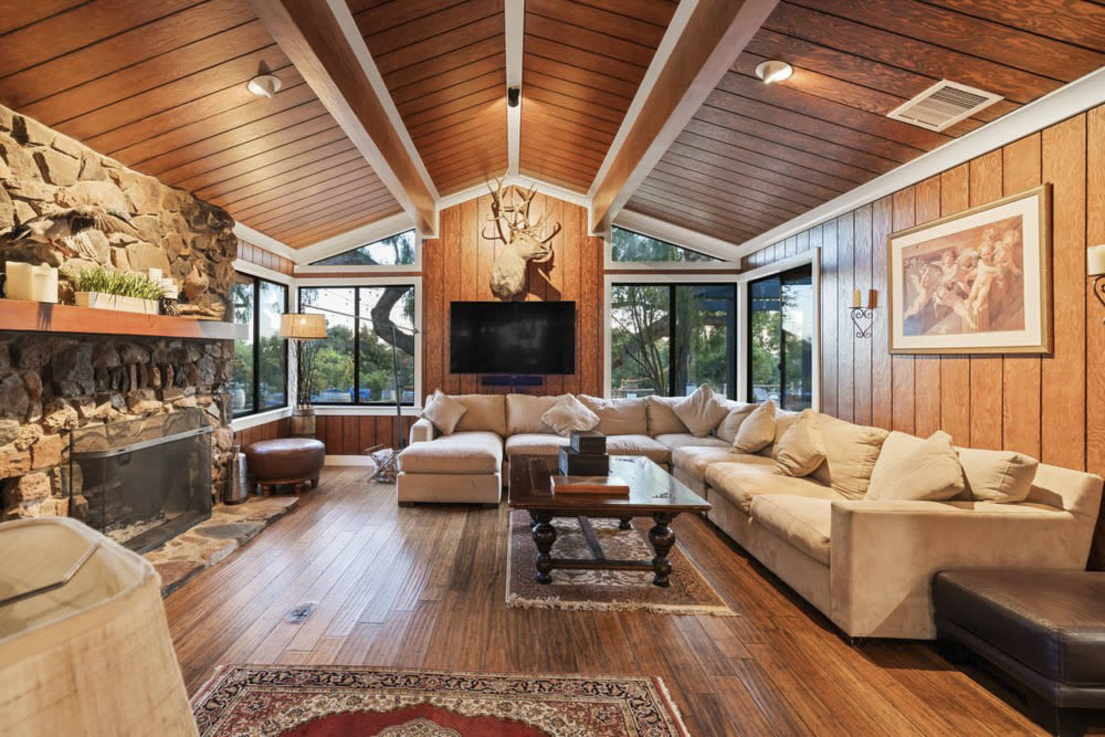 The image shows a cozy living room with wood-paneled walls, a stone fireplace, and a large sectional sofa arranged around a wooden coffee table.