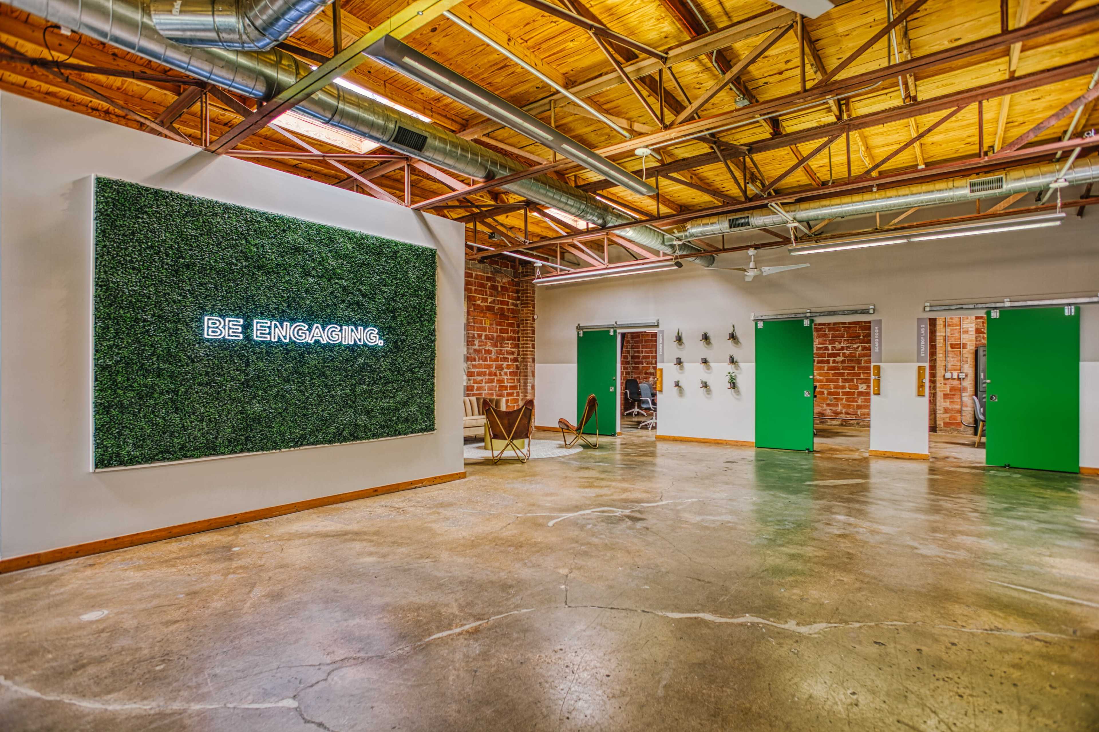 The image shows an industrial-style interior with a green wall featuring the phrase "BE ENGAGING" illuminated, contrasting against a concrete floor and exposed wooden beams.