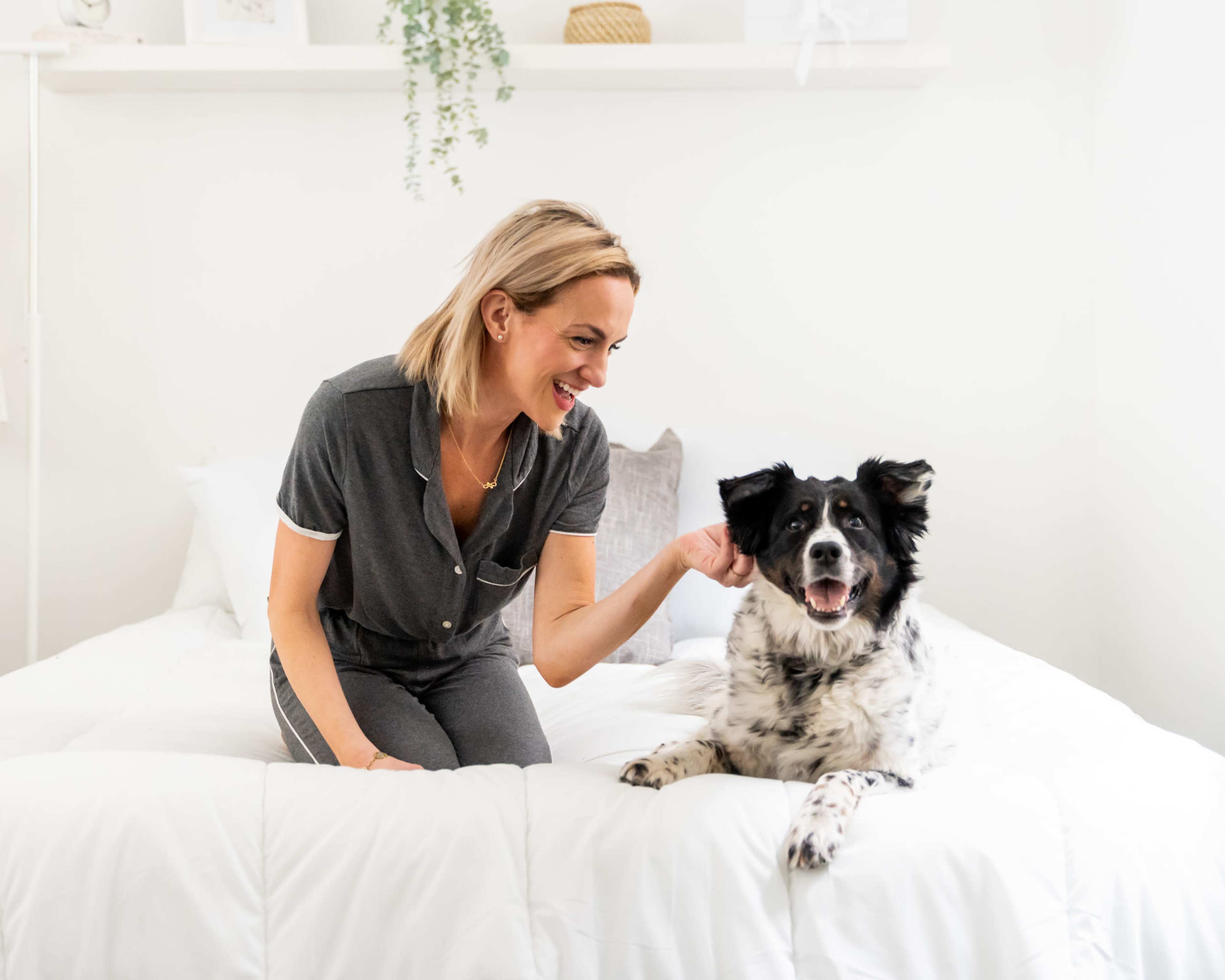 A woman sits on a bed with a dog, smiling while gently petting it in a bright, minimalist room.