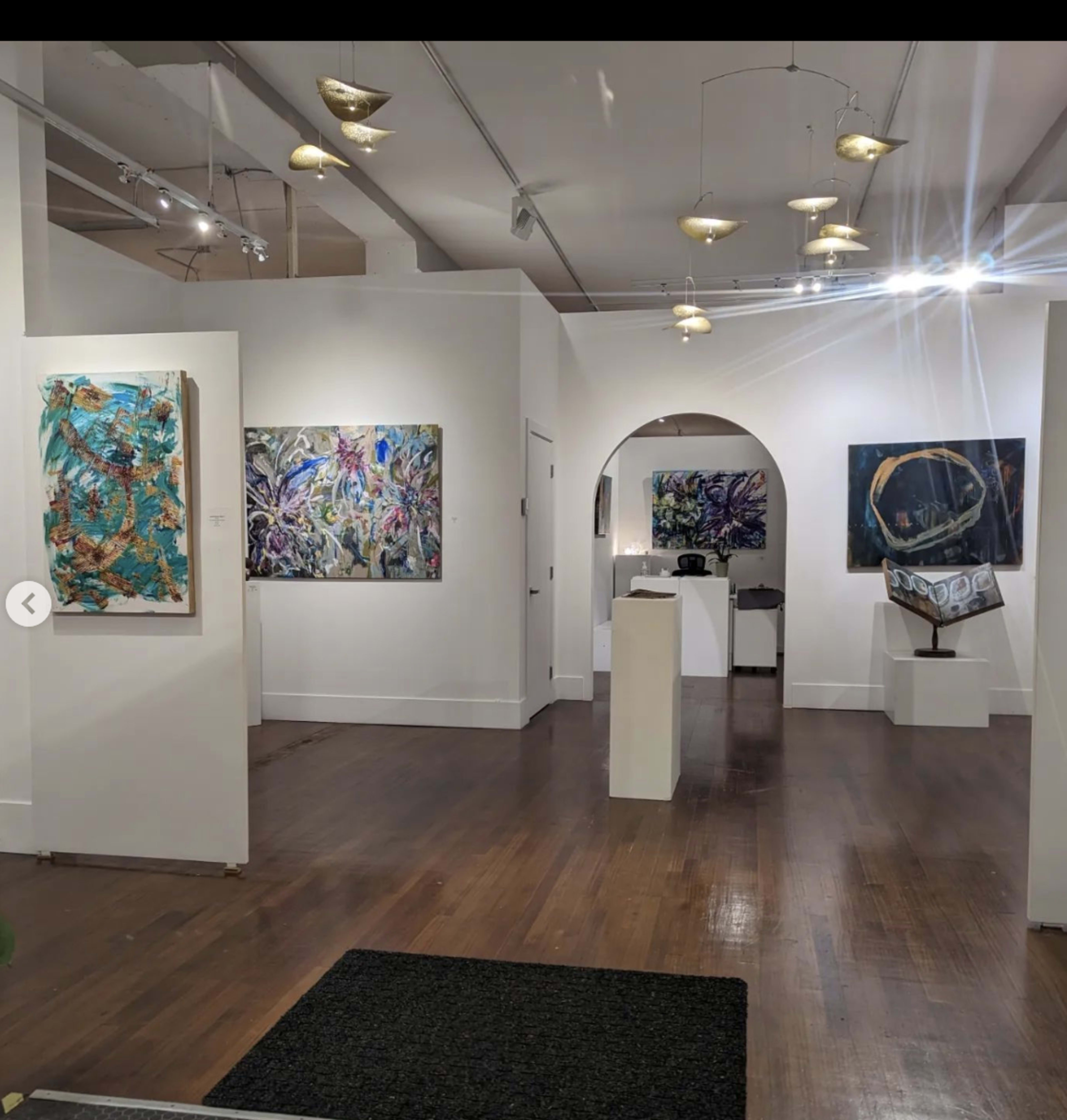 The image shows a modern art gallery with paintings displayed on the white walls and a sculpture on a pedestal in the center.