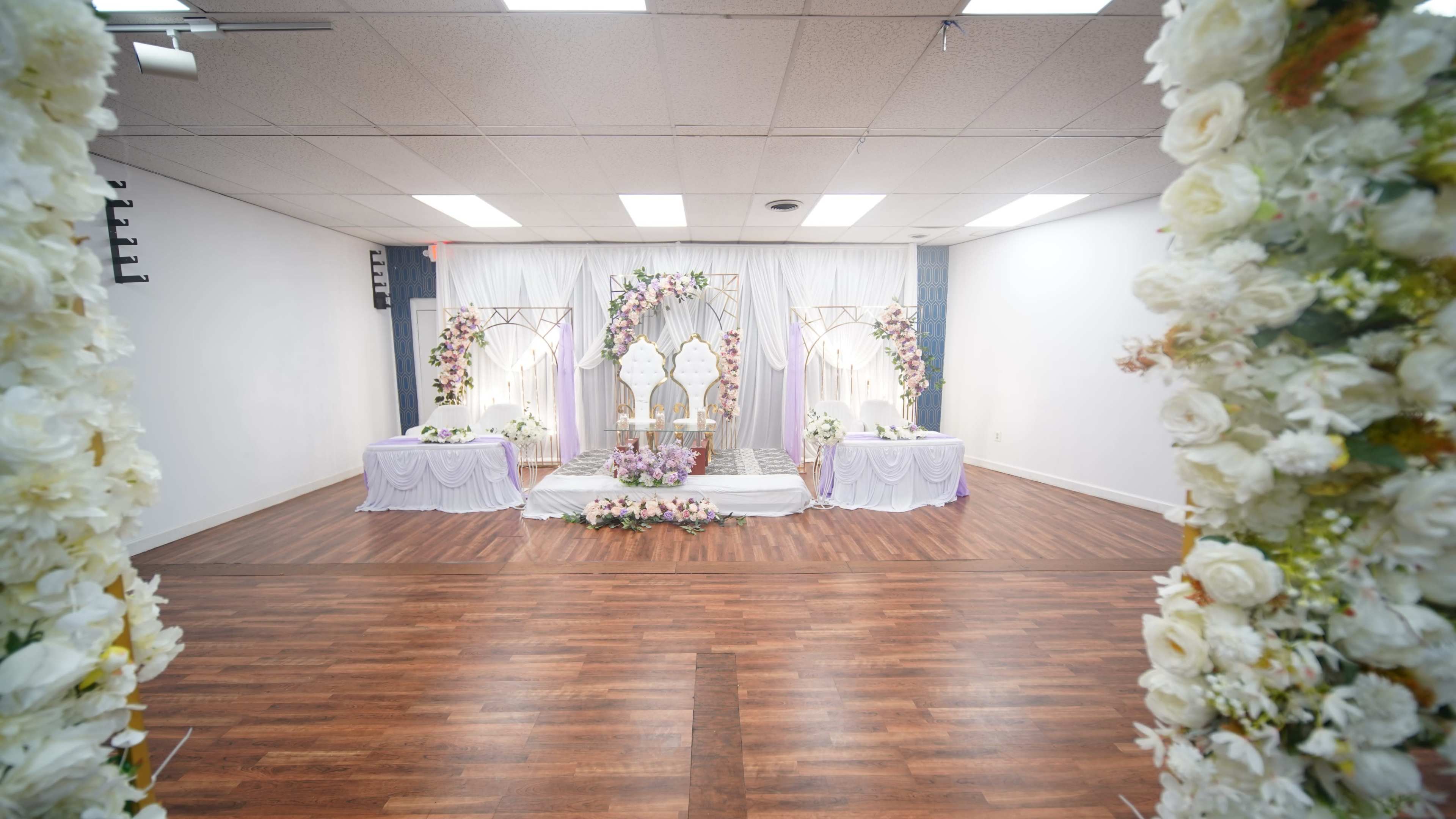 The image shows a decorated event space featuring a stage with floral arrangements, a central backdrop, and tables set up for a formal occasion.