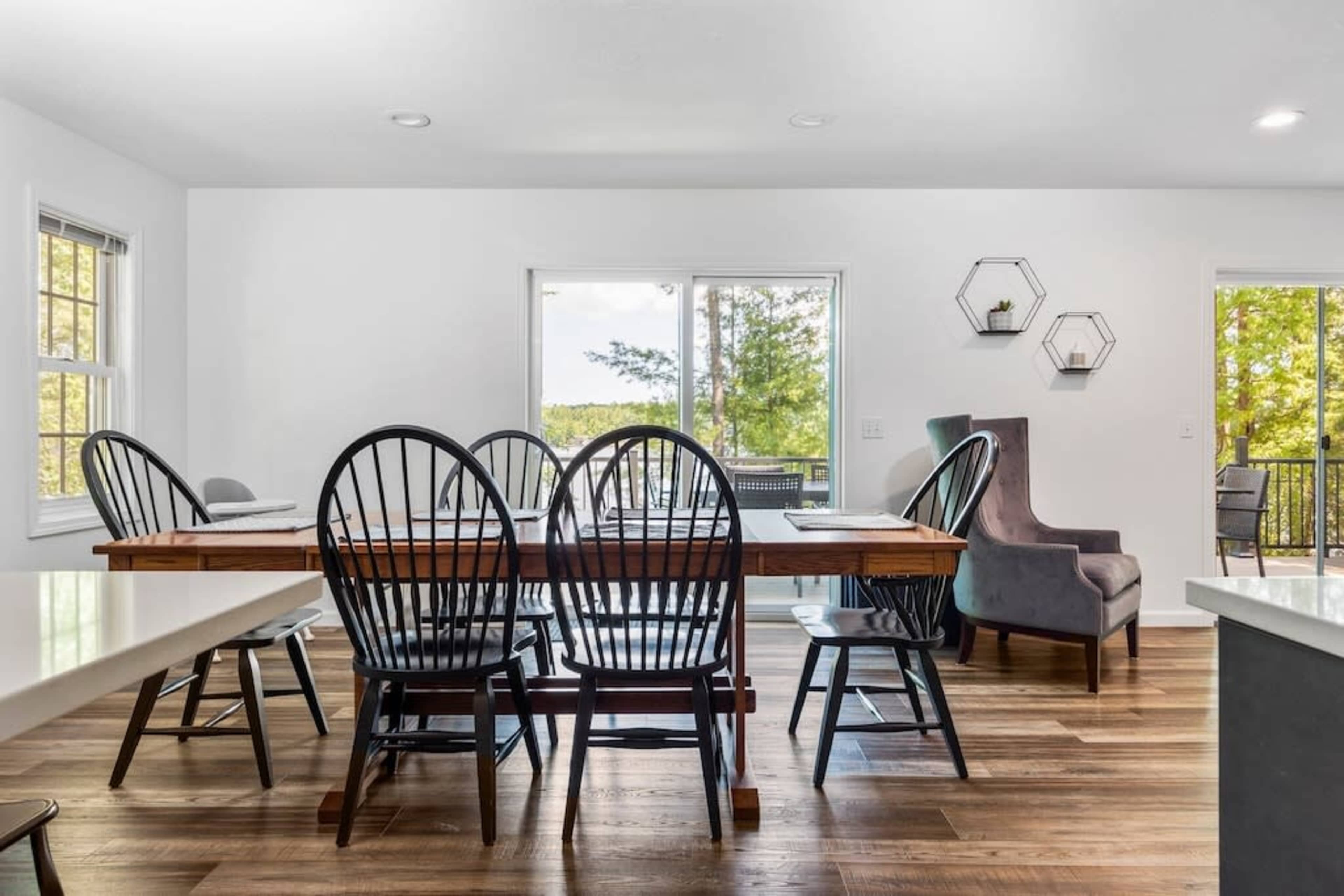 A wooden dining table with black chairs is placed in a well-lit room featuring large windows and minimalist decor.