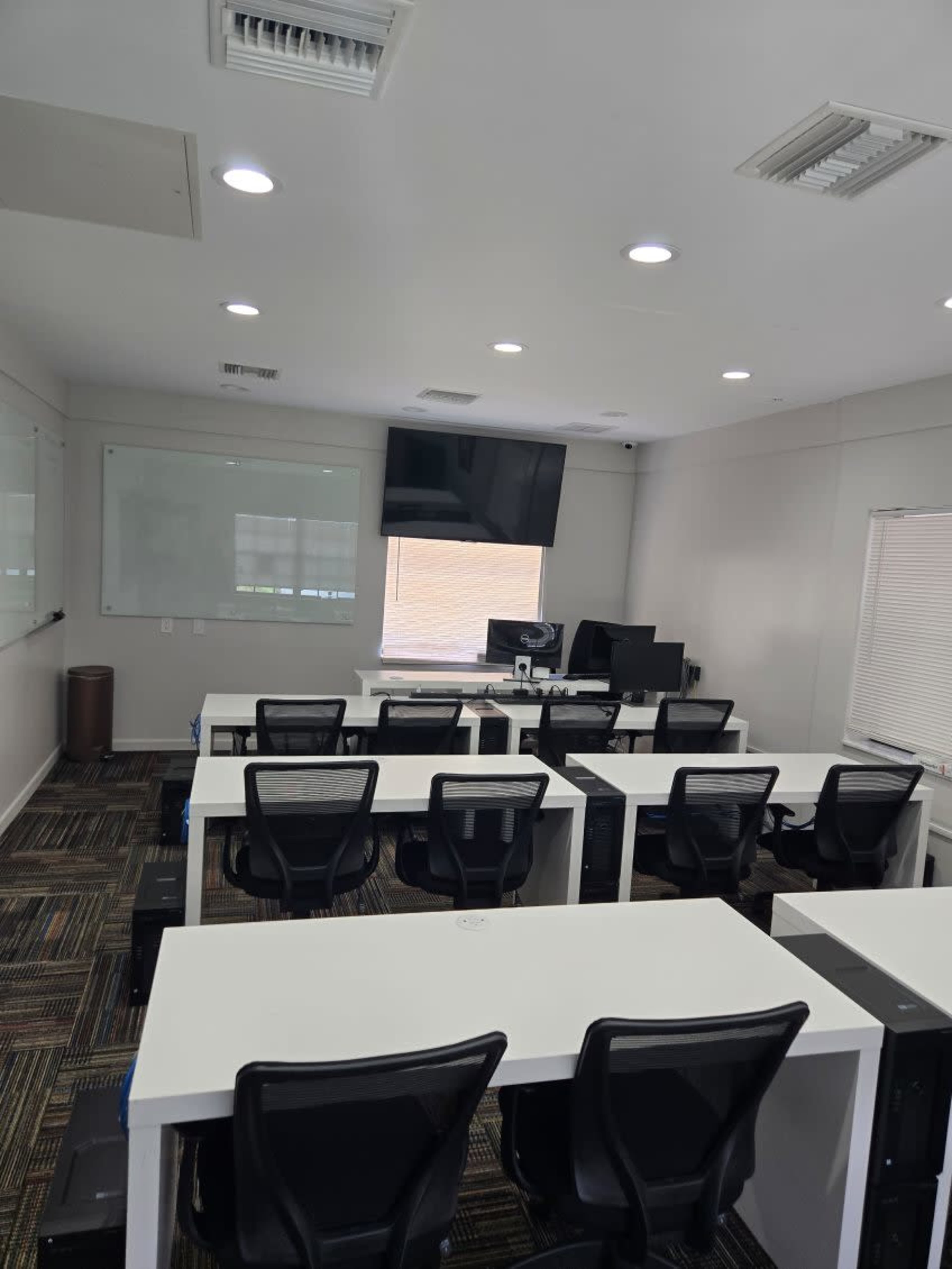 The image shows a modern classroom with rows of white desks and black chairs, featuring a large screen and computer setups in the front.