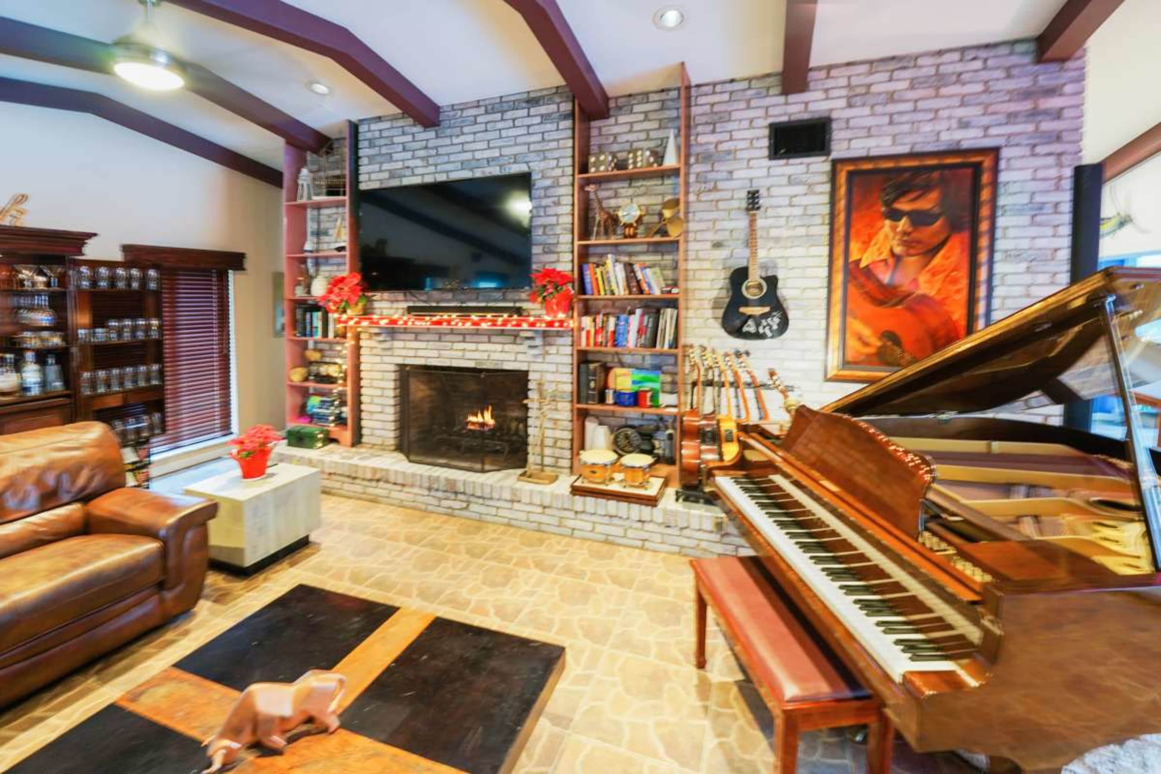 The image shows a cozy living room featuring a brick fireplace, a grand piano, bookshelves filled with various items, and a television mounted above the fireplace.