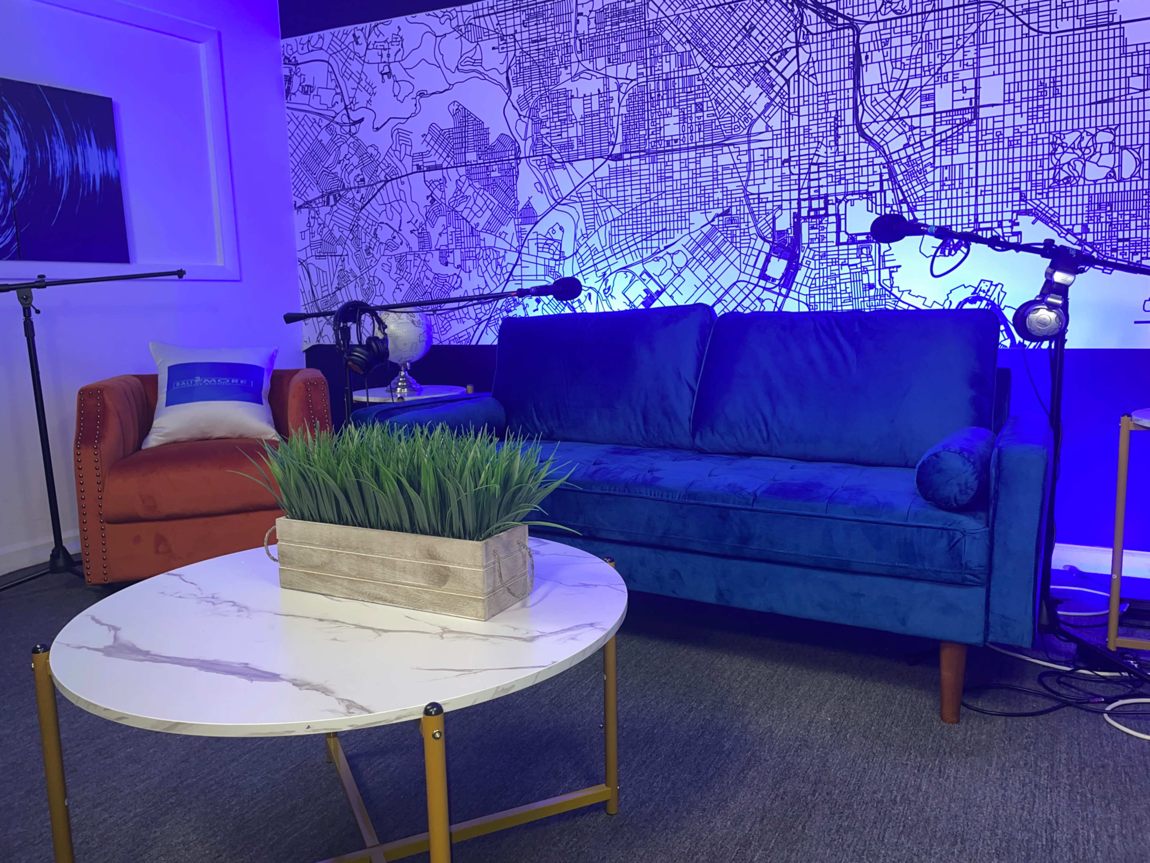The image shows a modern recording studio with a blue velvet sofa, an orange armchair, a round marble coffee table, and a wall featuring a large city map.