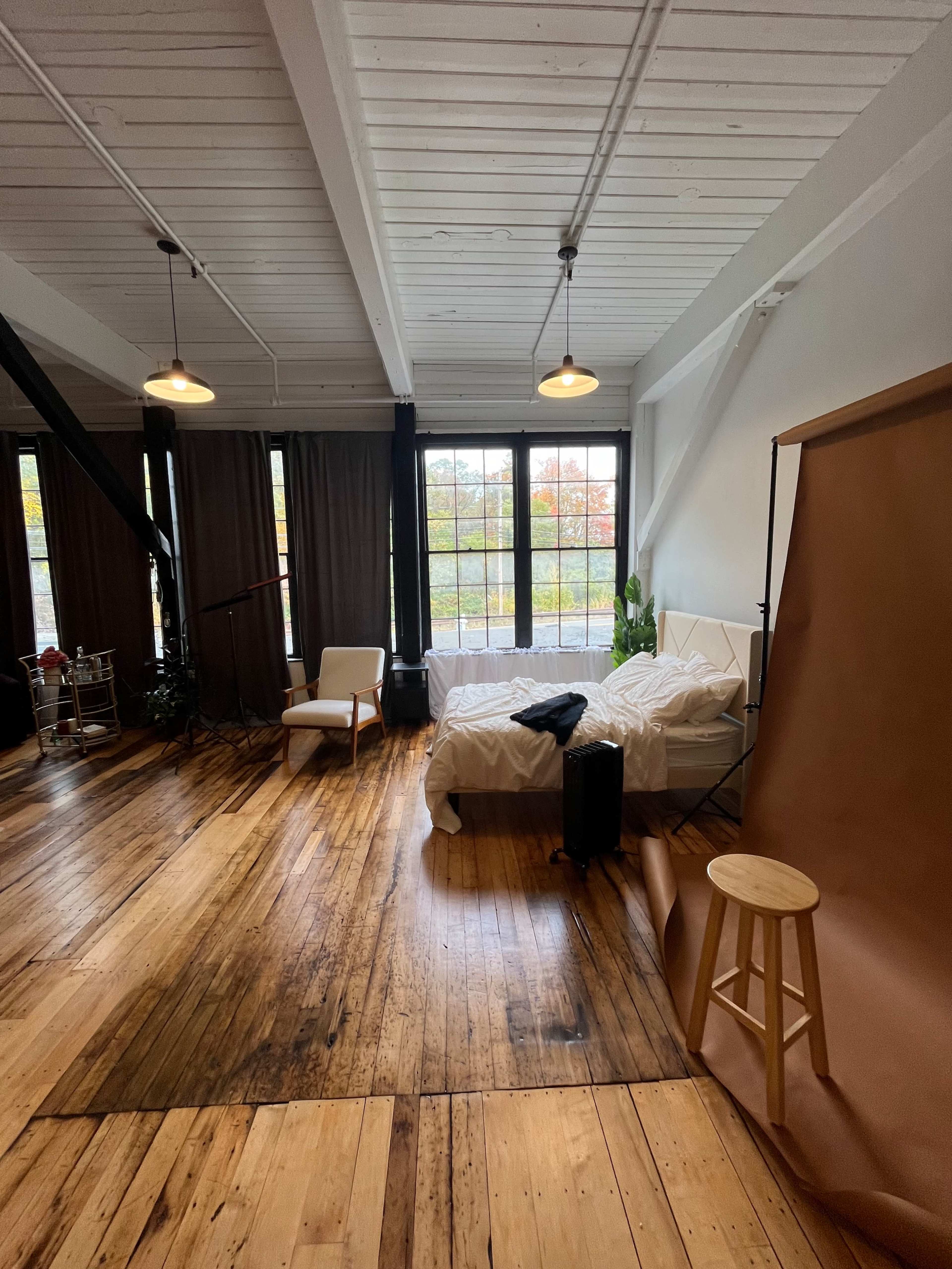 A spacious room with wooden floors, large windows, a white sofa with a rumpled bedding, and a simple stool near a brown backdrop.