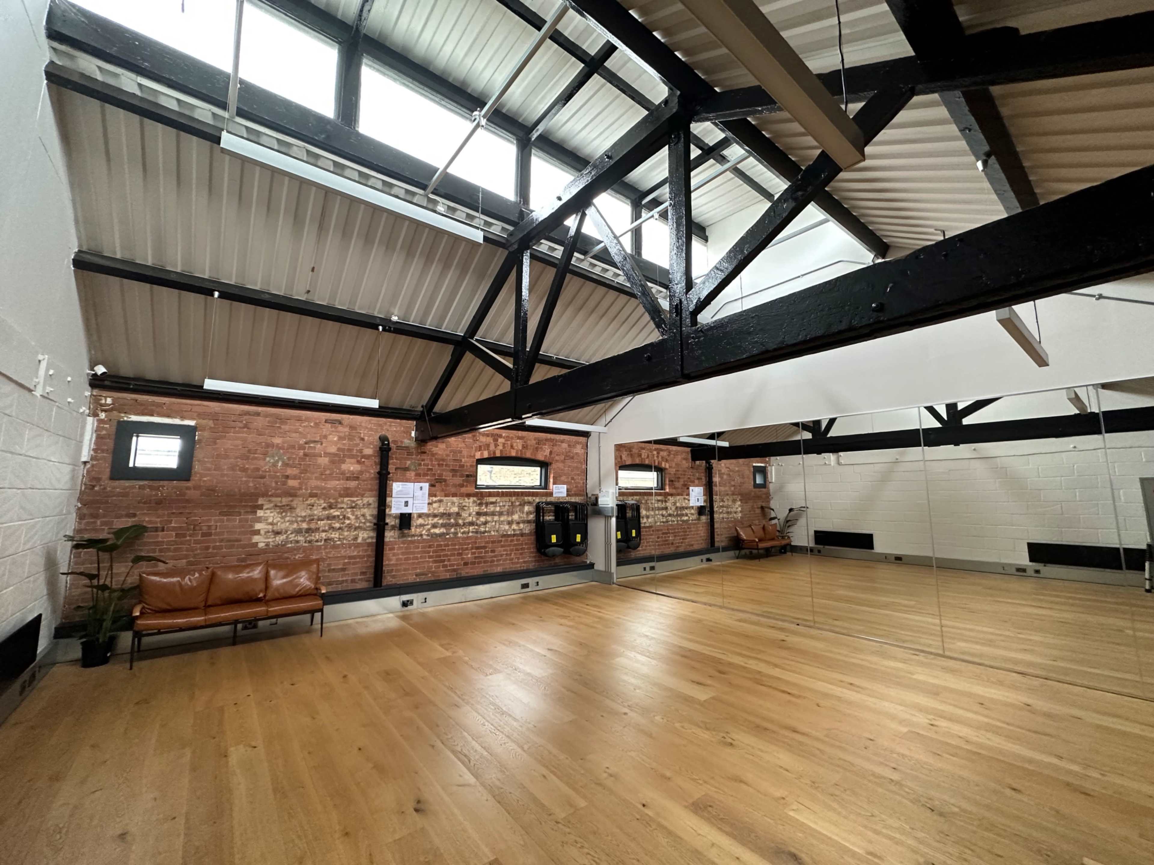 An interior of a spacious studio with wooden flooring, exposed brick walls, and high ceilings supported by black beams.