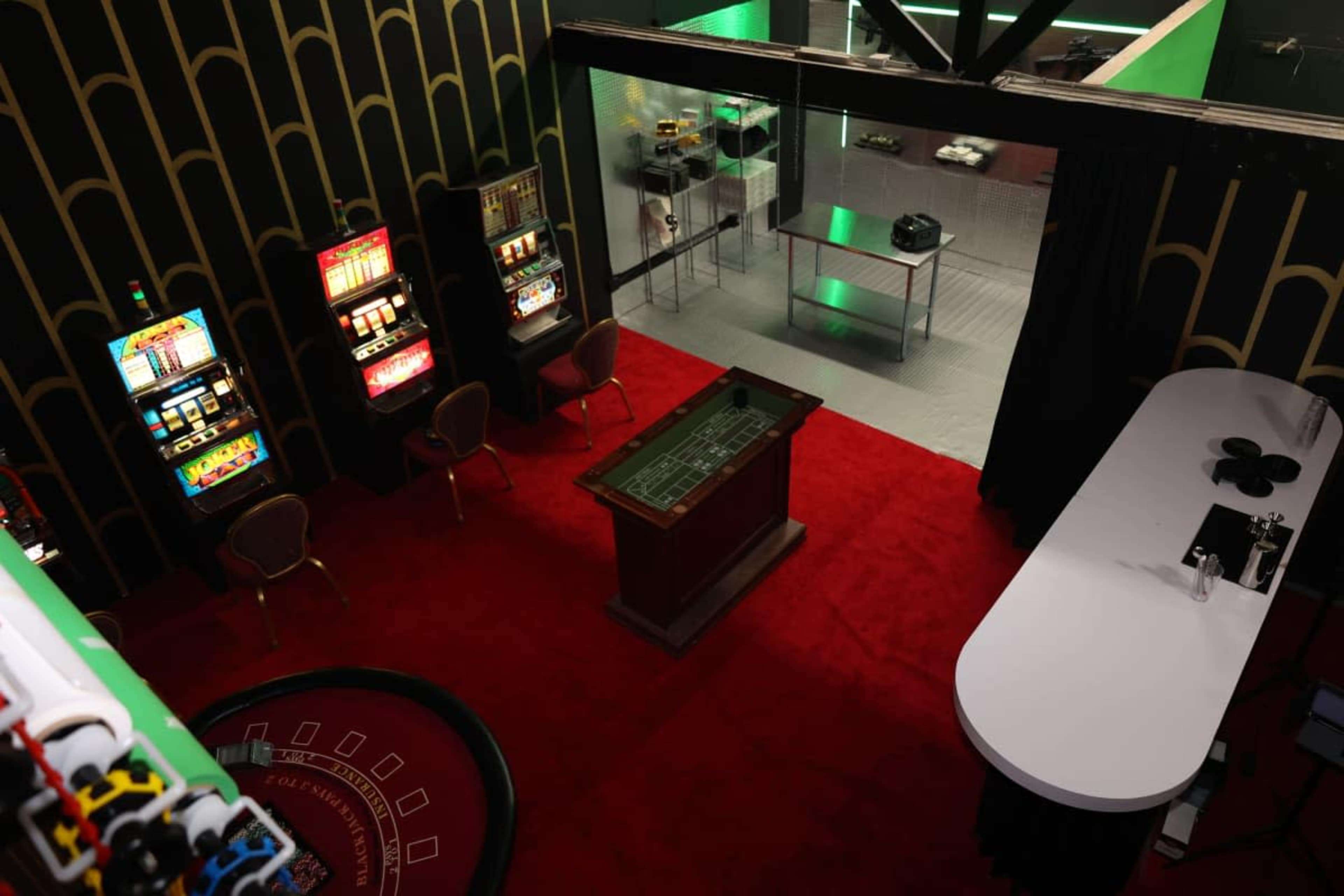 The image shows a gaming area with several slot machines, a roulette table, and a bar counter on a red carpeted floor.